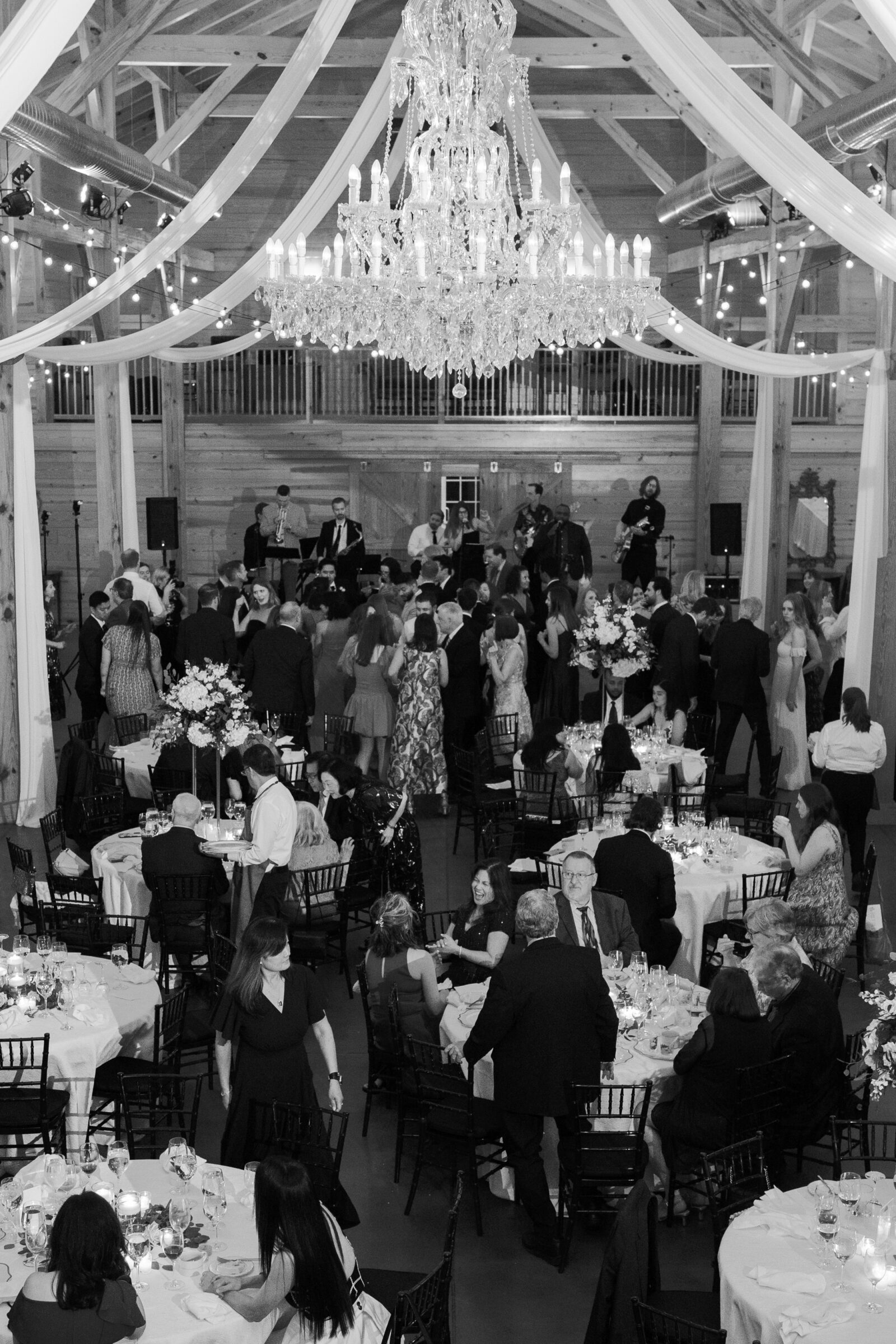 A black-and-white photo of the wedding reception at a barn venue. The guests are dancing and celebrating while a live band performs on stage. A grand chandelier hangs overhead, and floral arrangements decorate the tables. Some guests are seated at the tables while others are standing, enjoying the lively atmosphere.