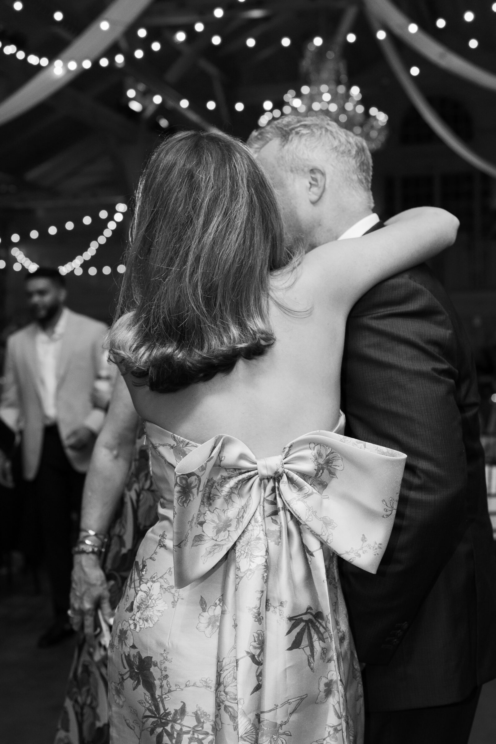 A black-and-white photo of a couple dancing during the wedding reception. The image focuses on the woman’s back, showcasing her floral dress with a large bow on the back. The man, in a suit, holds her as they dance, creating an intimate and romantic atmosphere. The venue is softly lit with string lights in the background.