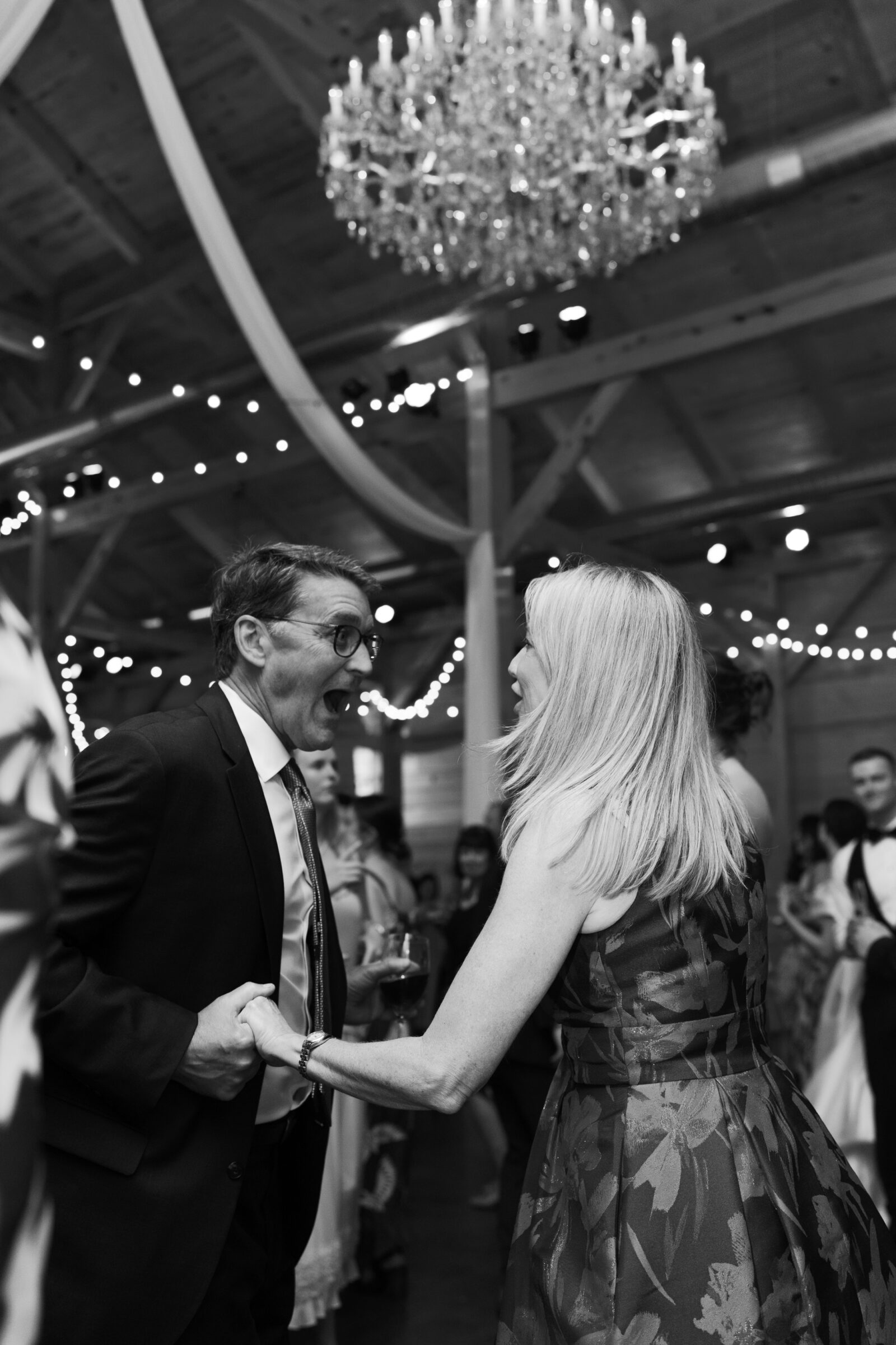 A black-and-white photo of two guests dancing and laughing at the wedding reception. The man is dressed in a suit and the woman is wearing a floral dress. They are holding hands while laughing and having a lively conversation, with a large chandelier overhead and string lights adding a festive atmosphere to the venue. The dance floor is filled with joyful people in the background.