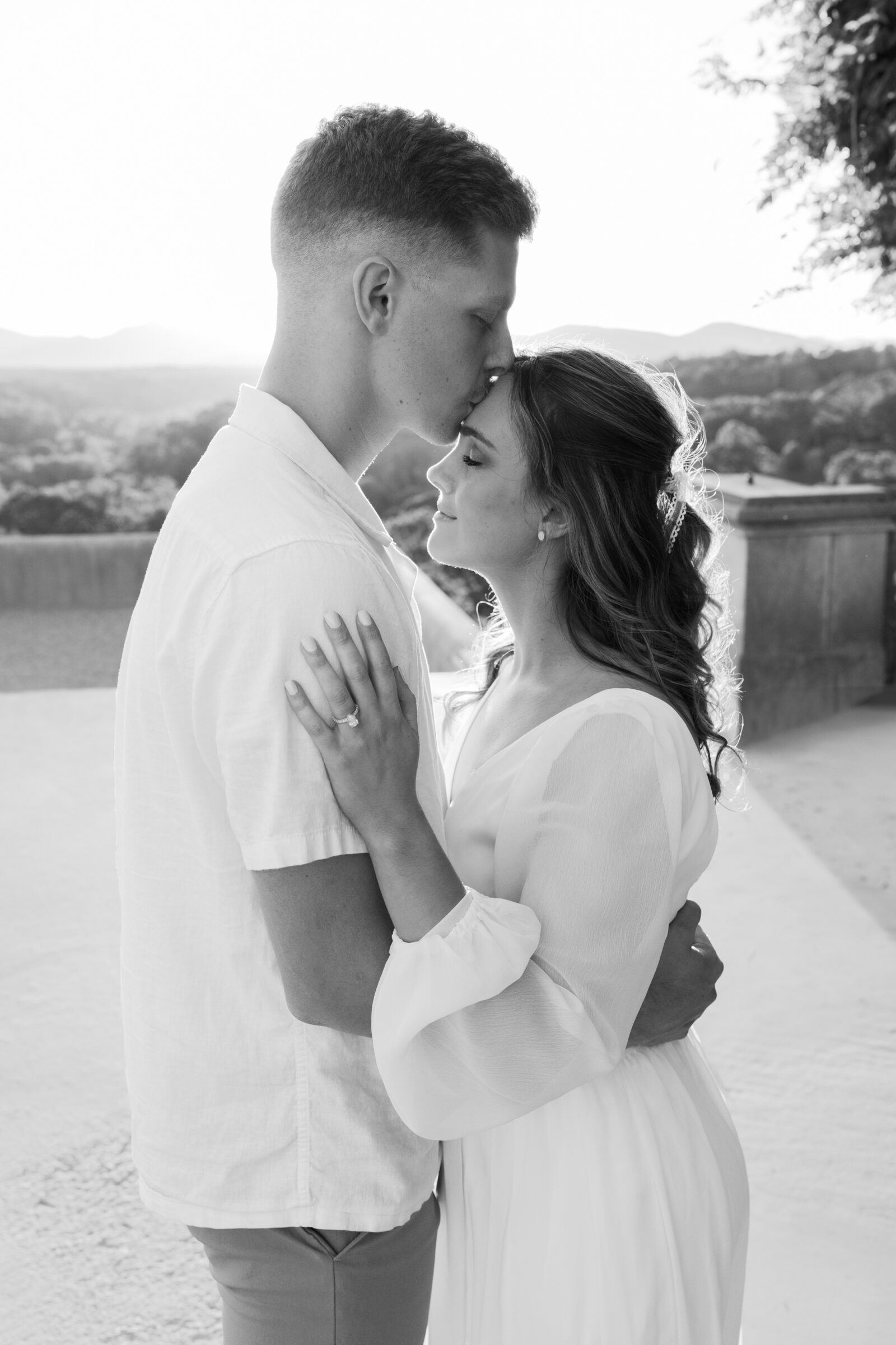 Engagement Photos at the Biltmore Estate | black and white image of guy kissing girl's forehead on terrace with Appalachian mountains in the background