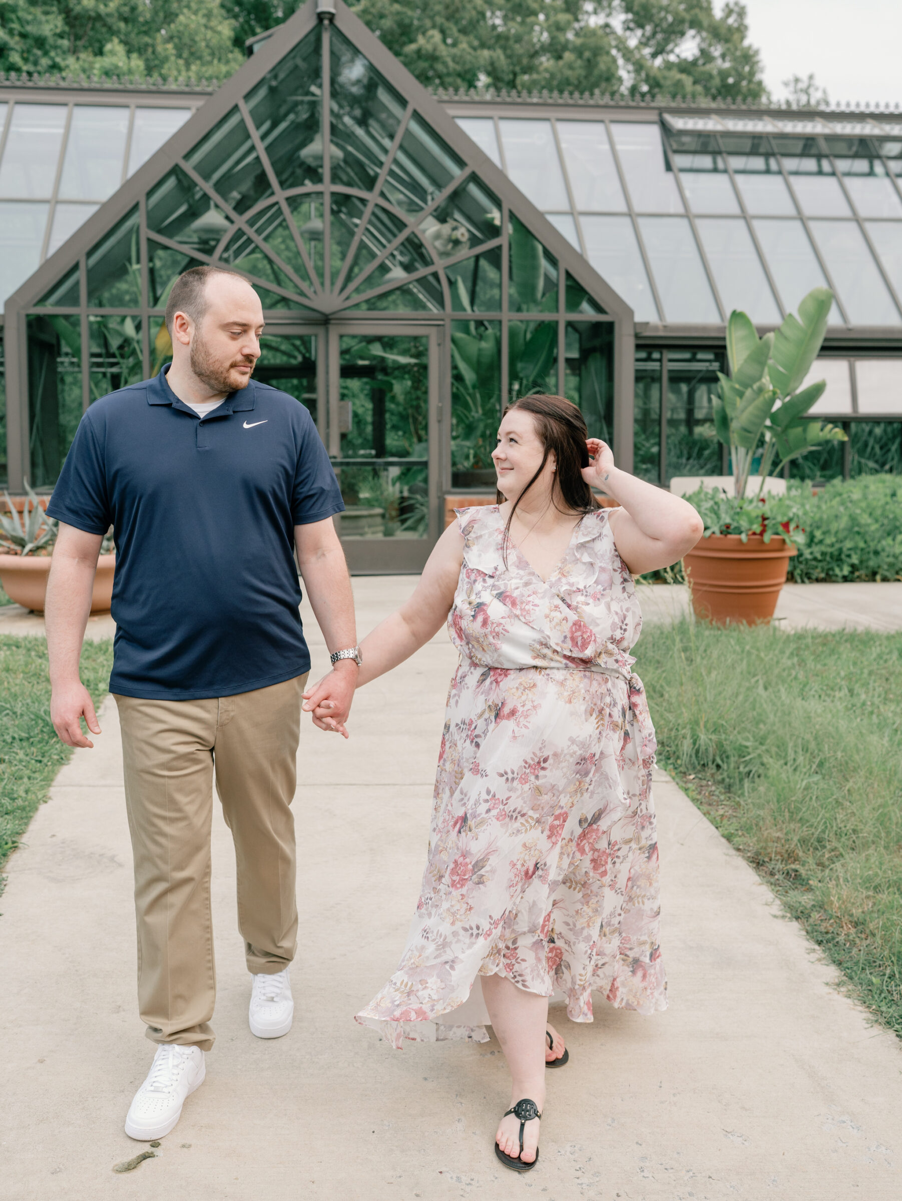 Meadowlark Botanical Gardens Engagement Photos | girl in floral dress holds her hair back as she walks hand-in-hand with her fiance in a navy blue polo in front of the Meadowlark Botanical Gardens greenhouse