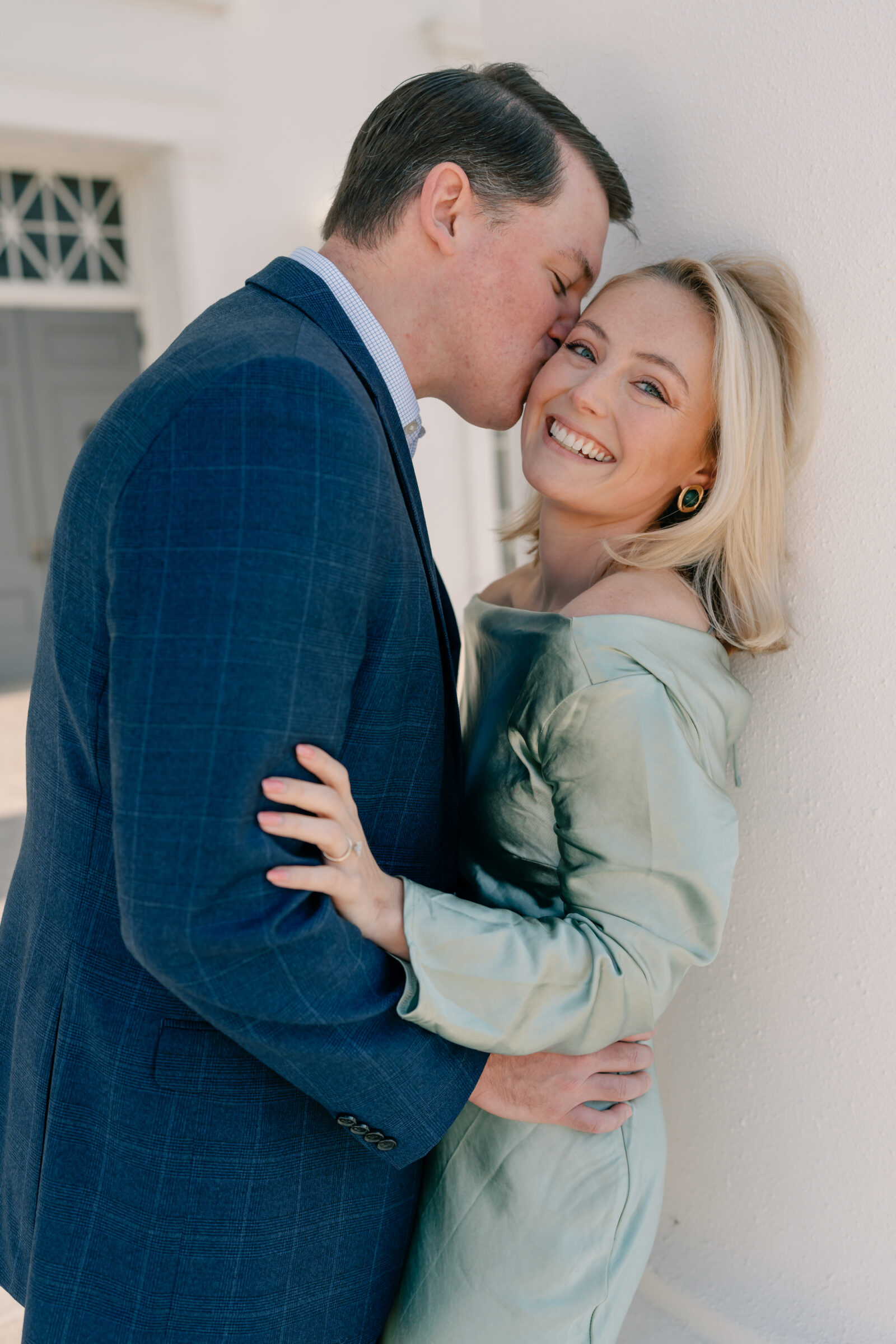 Richmond Virginia Engagement Photographer | newly engaged couple in green dress and blue suit lean against white column of the Virginia Capitol Building. The groom-to-be kisses his future wife on the cheek as she giggles