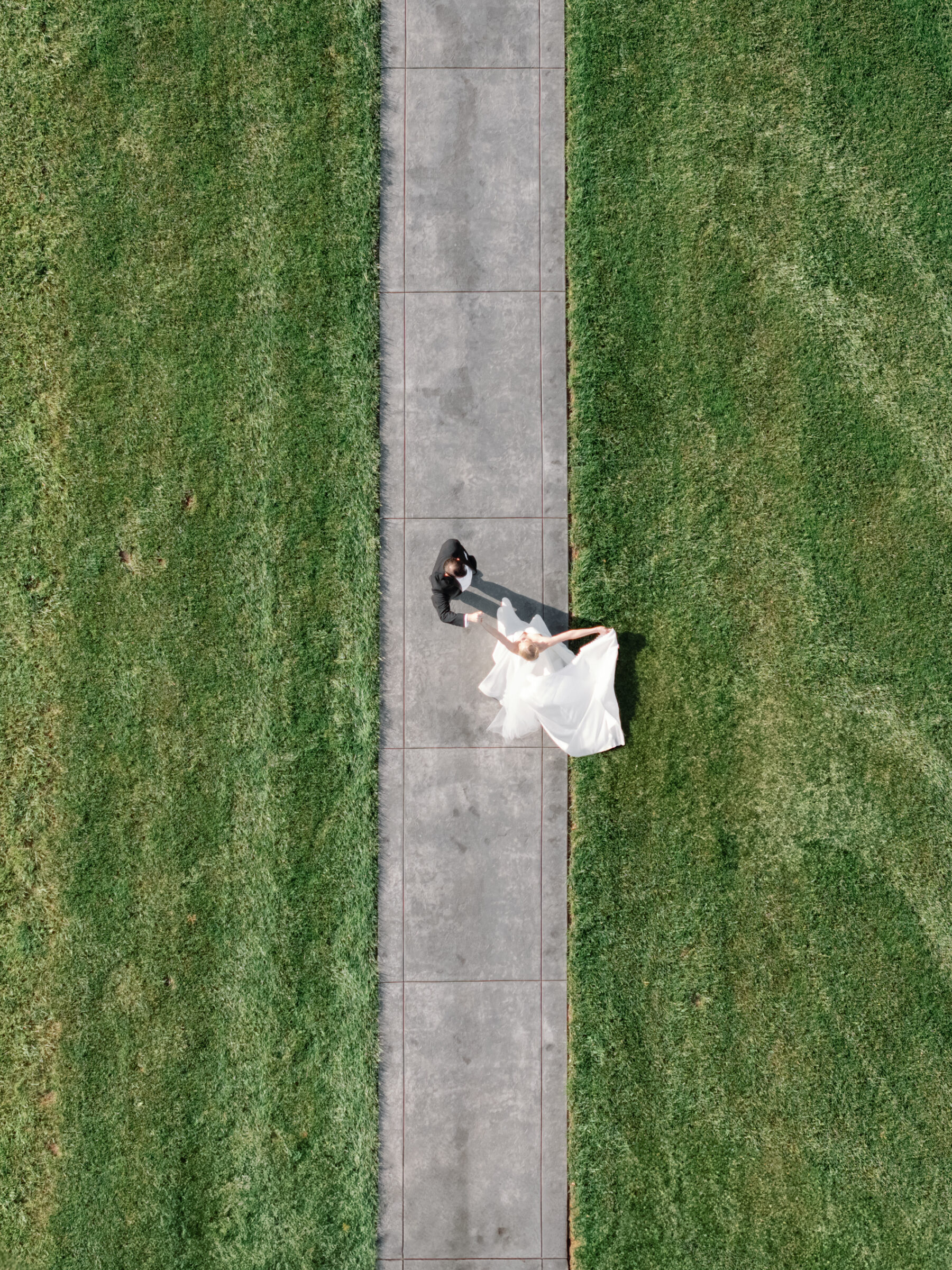 A higher drone shot looking straight down at the bride and groom on the same central path. Their bodies form abstract shapes—a white circle of gown and a dark figure—amid the broad expanse of turf, emphasizing the symmetry of the scene and the elegance of their movement.