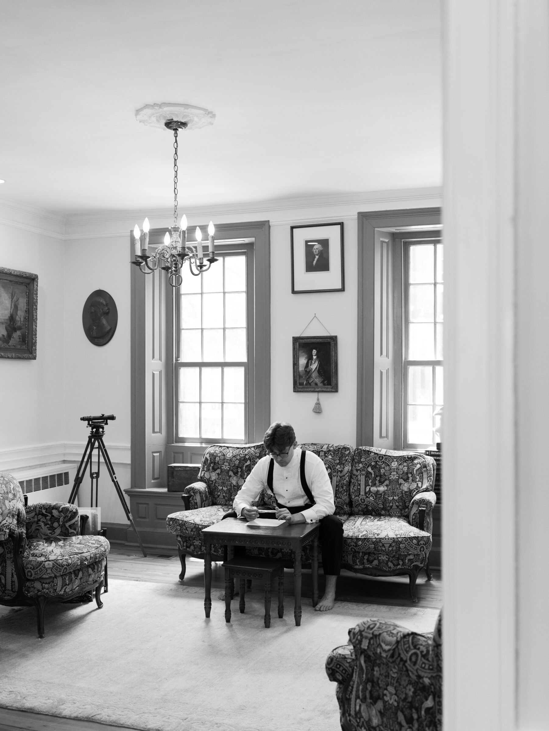 Alexandria Wedding Photographer captures the groom seated barefoot on a vintage sofa in a historic parlor, thoughtfully reading a handwritten note.