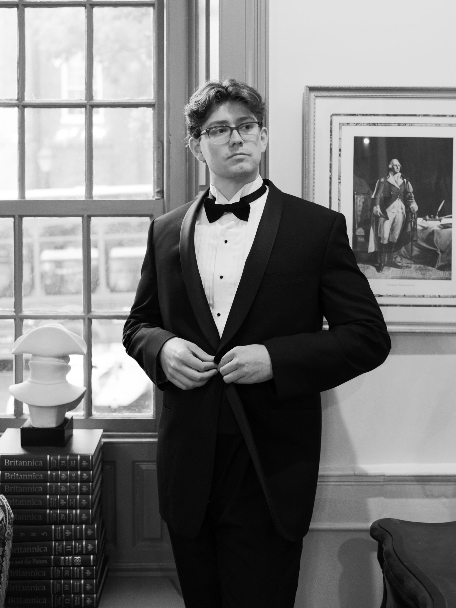 Groom adjusts his tuxedo jacket by a sunlit window in a historic Old Town Alexandria parlor, exuding refined, timeless elegance.