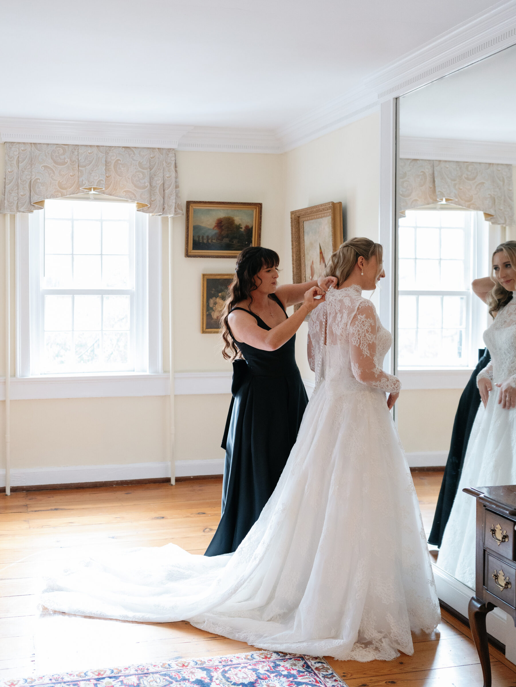 Alexandria Wedding Photographer frames a bridesmaid fastening the bride’s lace gown buttons as she admires her reflection in a sunlit room.