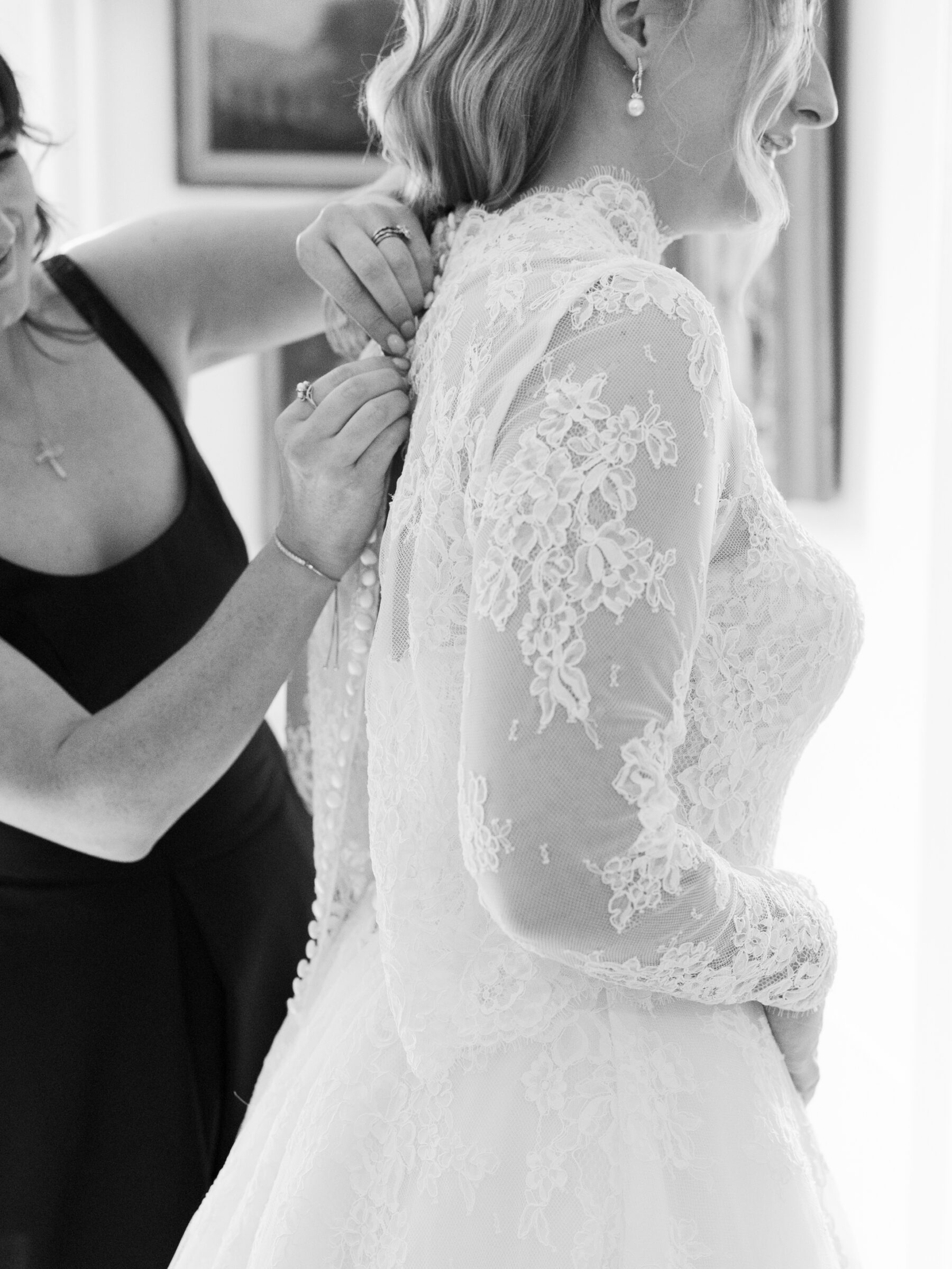 Alexandria Wedding Photographer captures a bride in a delicate lace gown as her bridesmaid fastens the back buttons in soft, natural light.