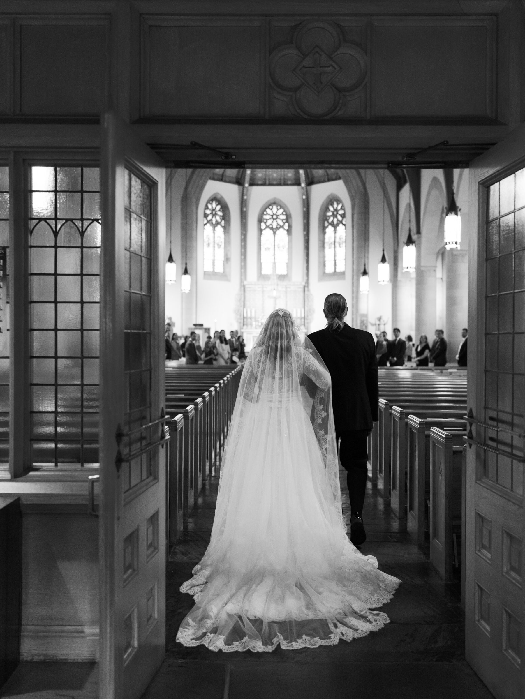 The bride and groom walk hand in hand through the open doors of St. Rita’s Catholic Church, their long lace train trailing over stone floors toward a sunlit altar.