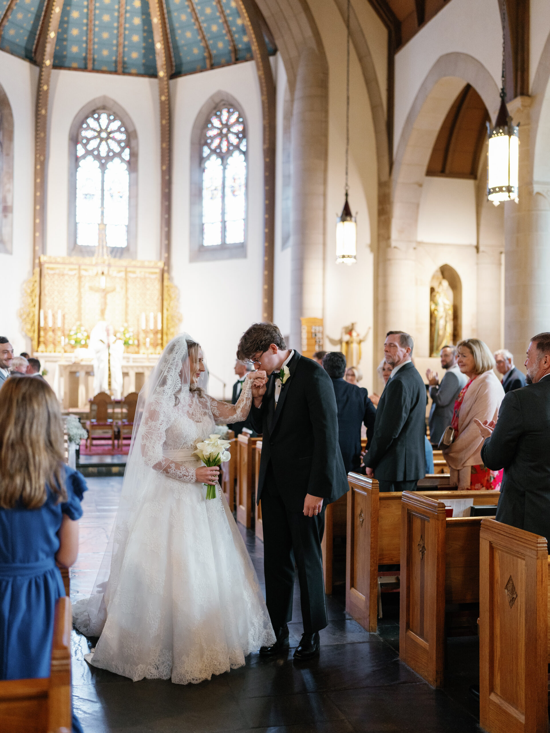 A groom’s gentle kiss on his bride’s hand in the aisle of St. Rita’s Catholic Church, showcasing the timeless elegance of an Alexandria Wedding Photographer’s work.