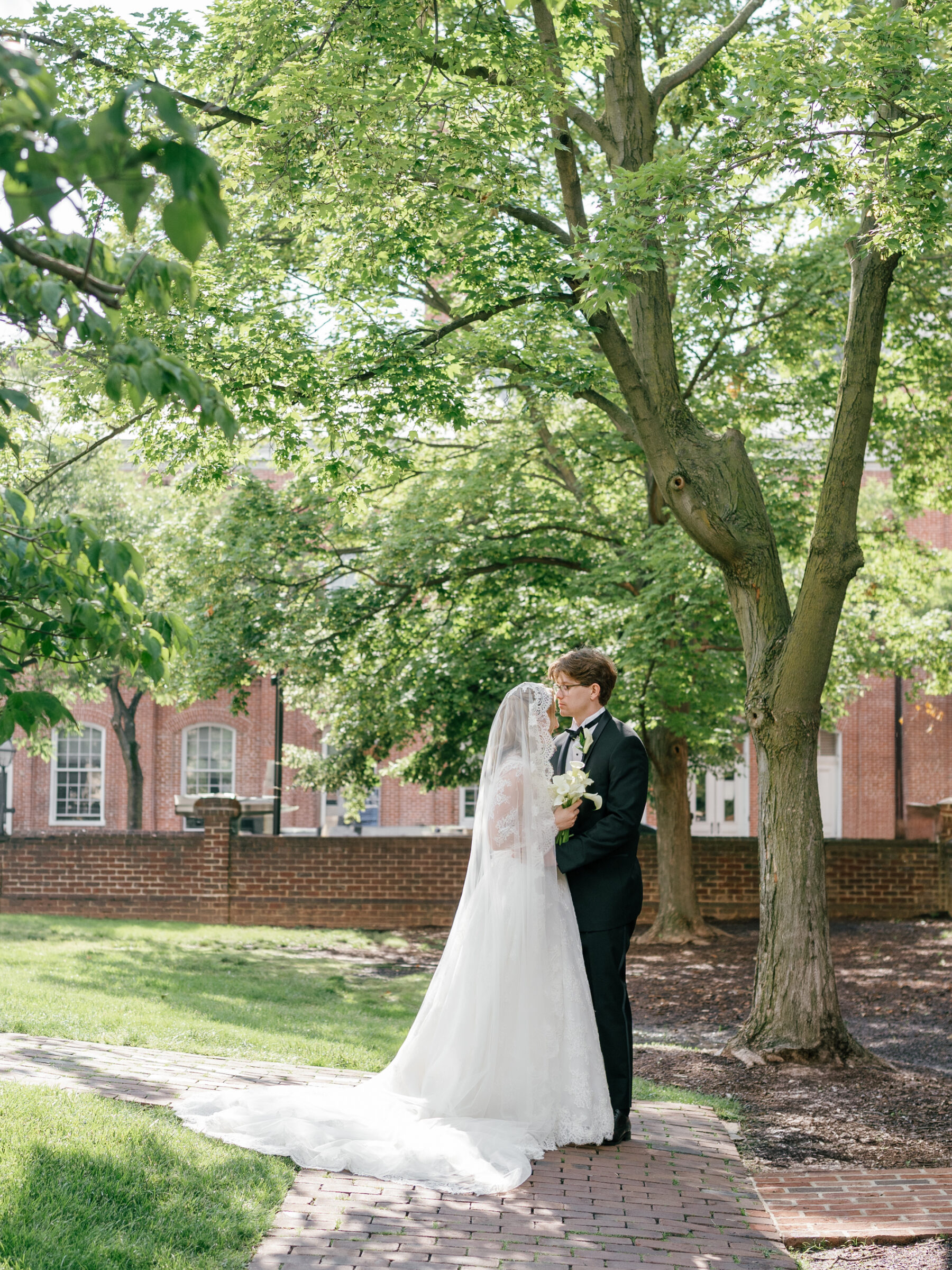Bride and groom share a tender moment beneath leafy branches at Carlyle House, the bride holding a bouquet of calla lilies as dappled sunlight filters through the trees.