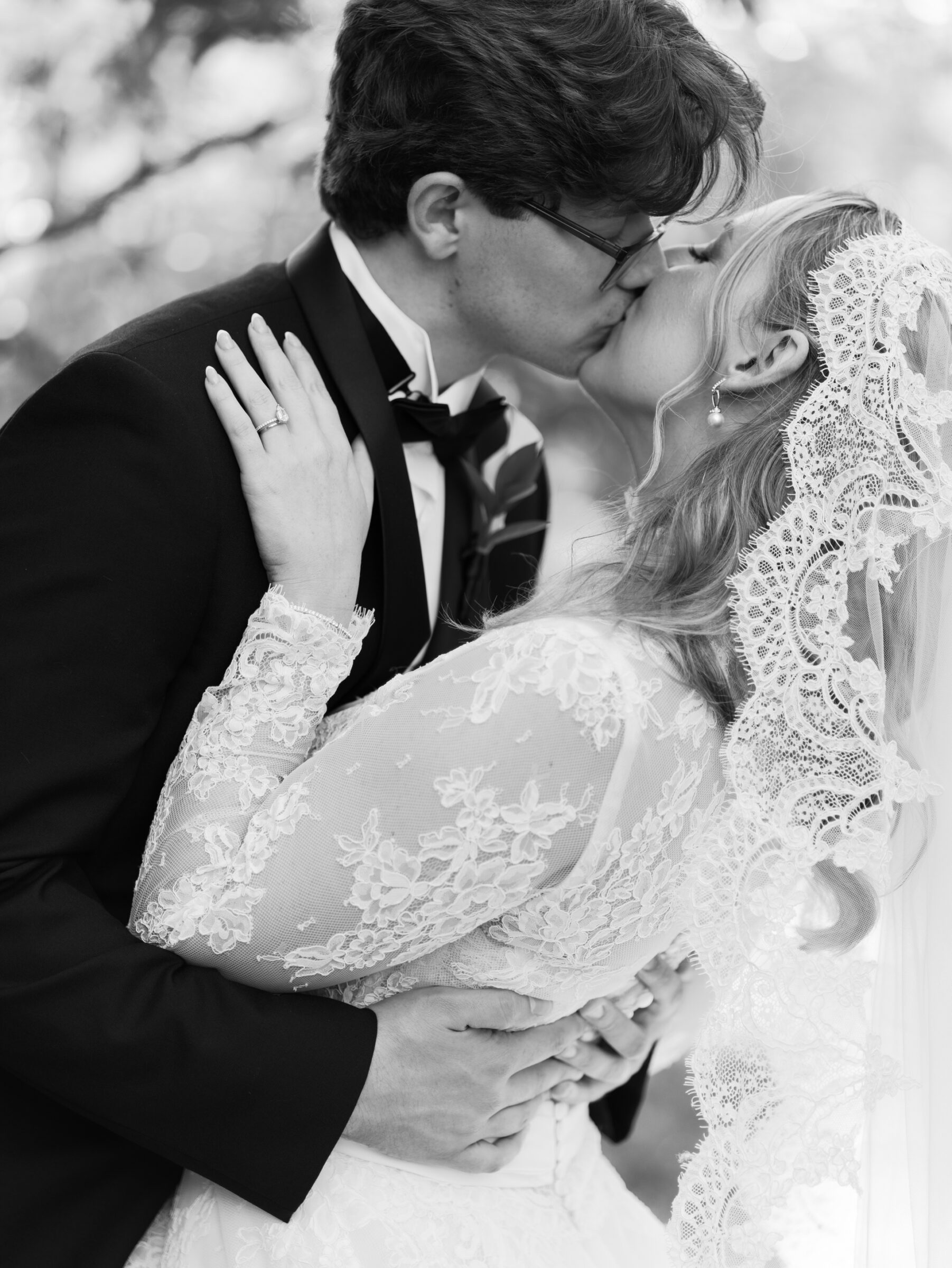 Bride and groom share a romantic dip and kiss beneath a leafy canopy, her lace veil cascading around them as he holds her gently by the waist.
