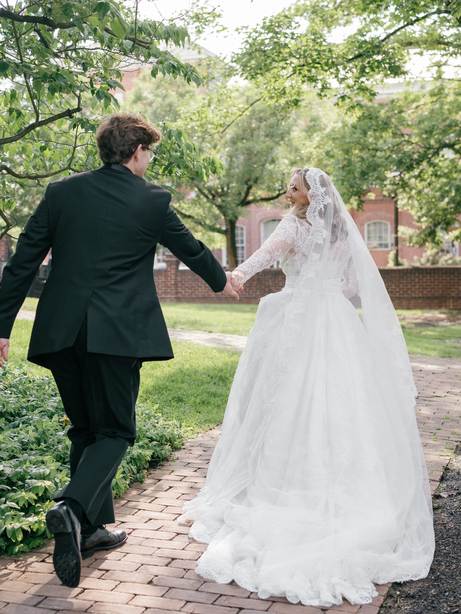 Bride and groom hand in hand walking along a sun‑dappled brick path beneath leafy trees at Carlyle House, captured by an Alexandria Wedding Photographer.