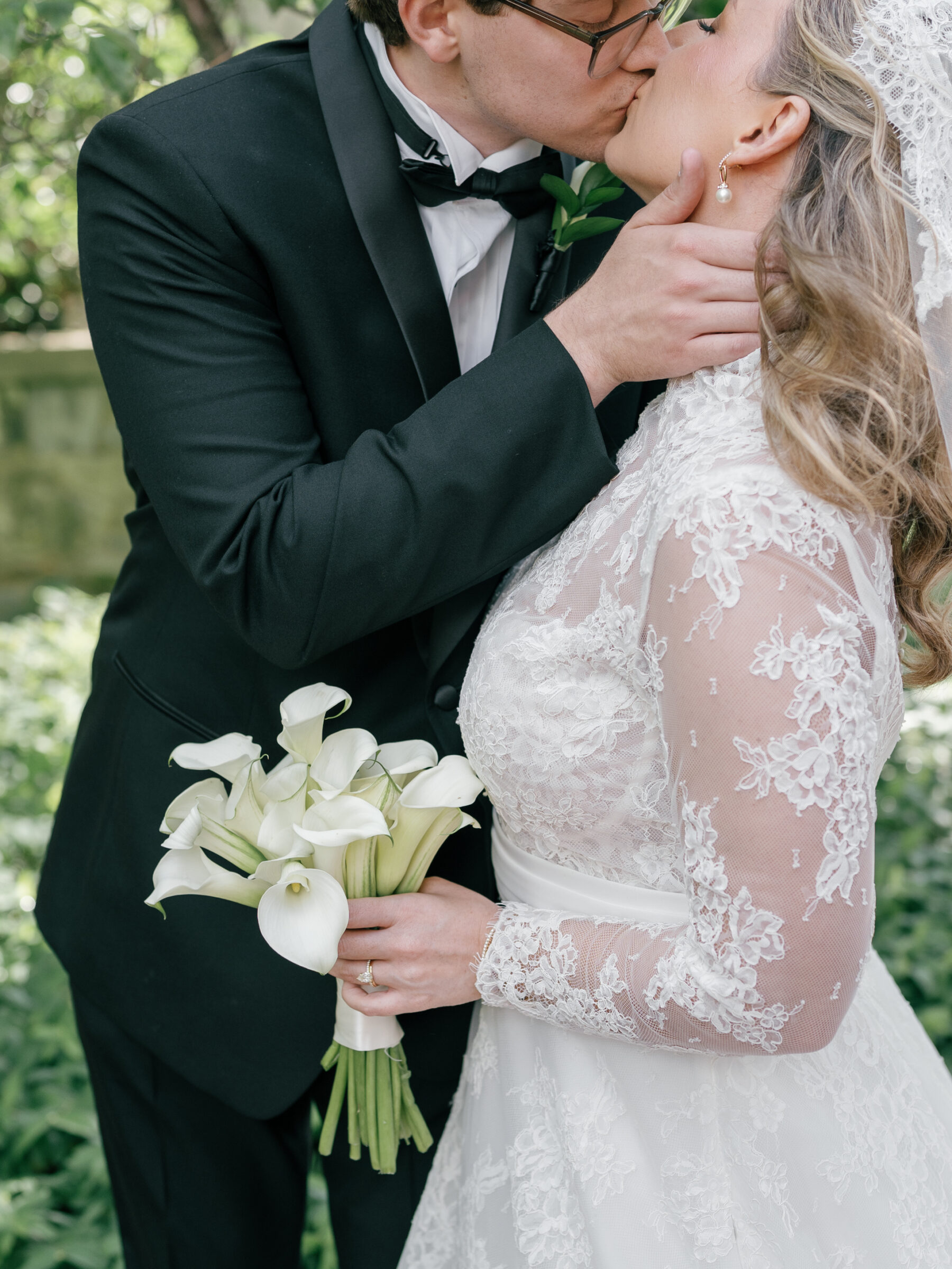Groom in a black tuxedo gently kisses the bride’s hand as she holds a bouquet of white calla lilies beneath leafy green trees at Carlyle House.