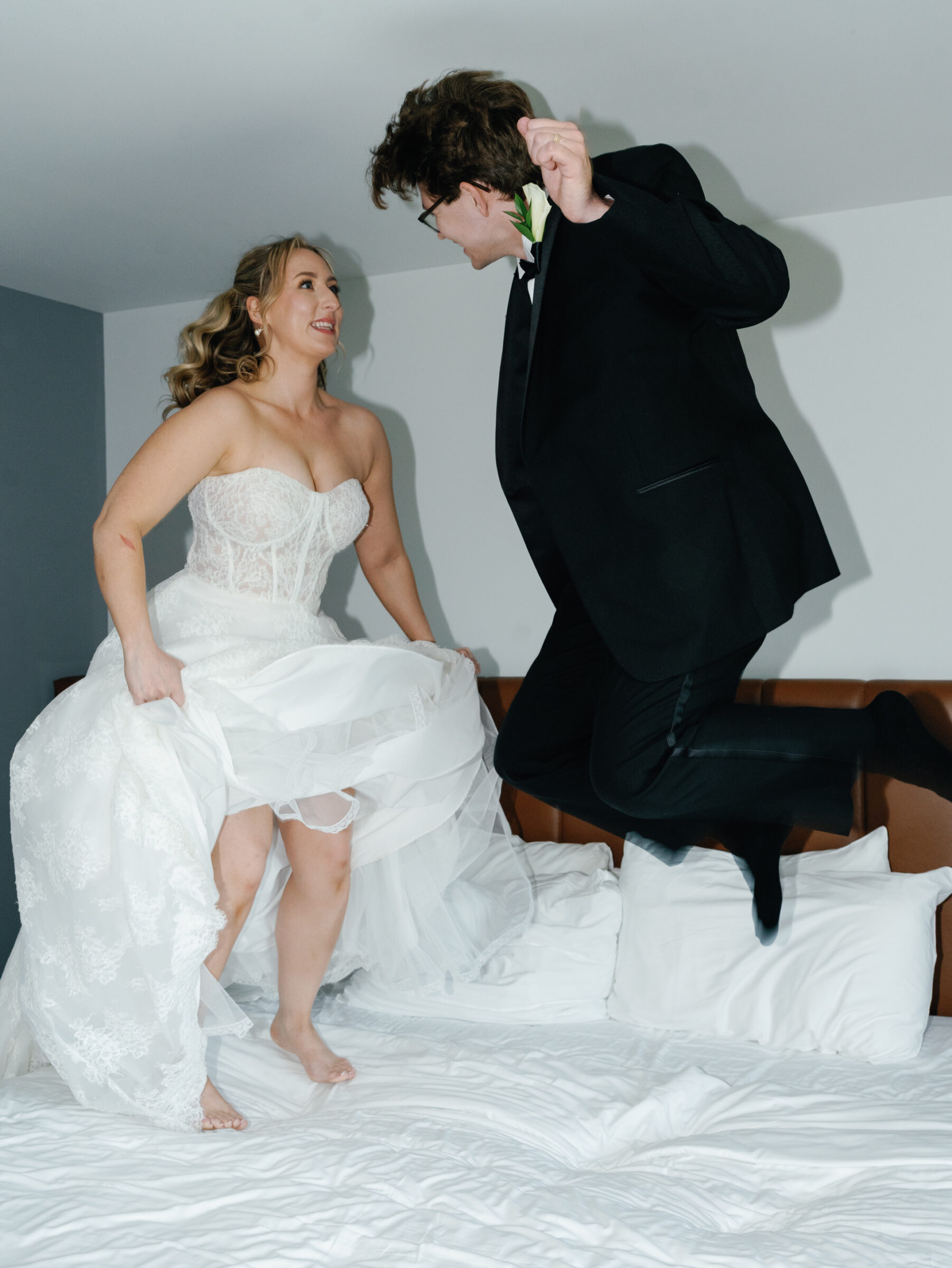 Bride in a lace gown and groom in a tuxedo laugh as they joyfully jump together on a crisp white bed in their honeymoon suite.