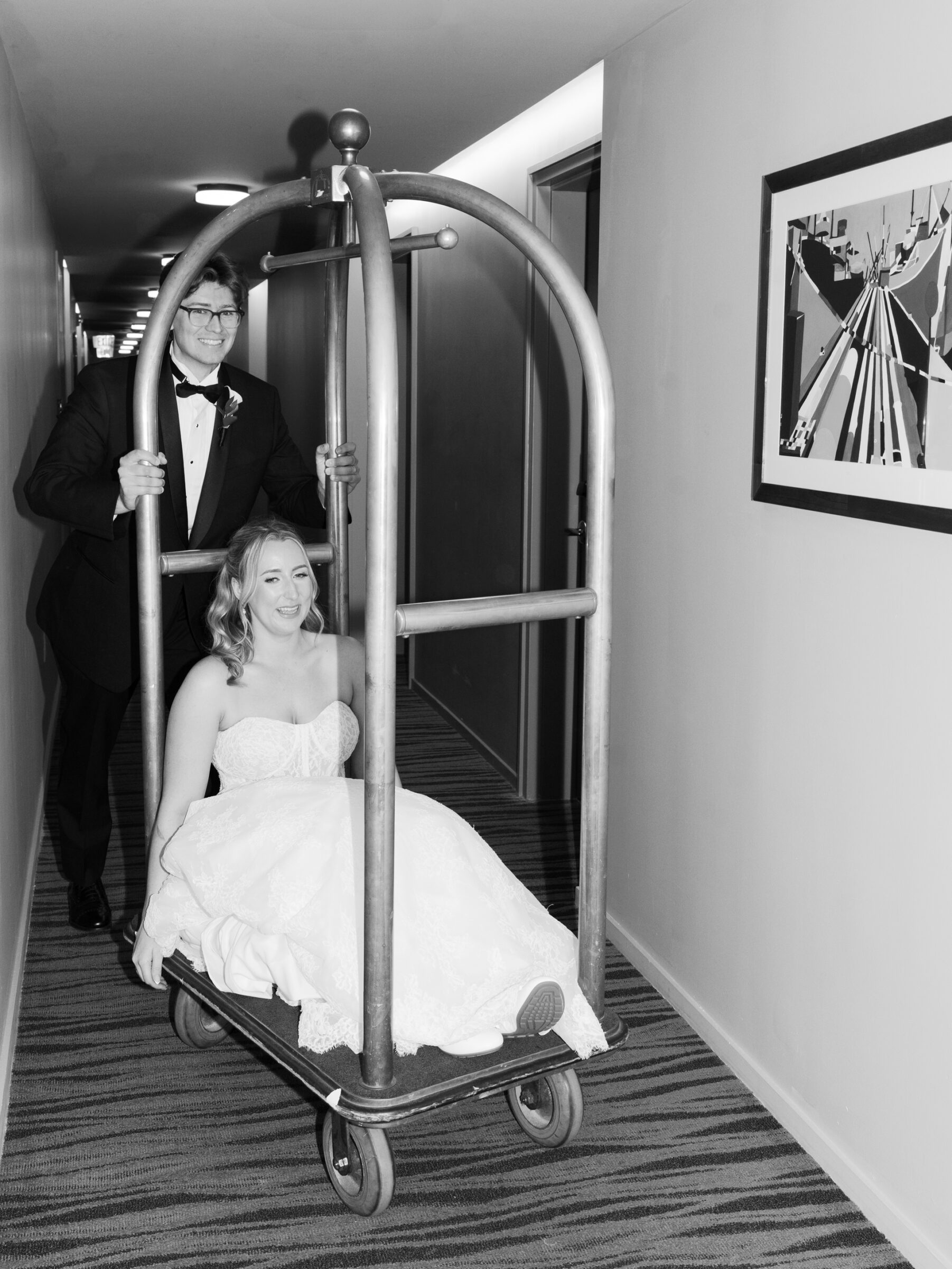 Bride laughs as she sits on a hotel luggage cart while her groom playfully pushes her down a hallway, captured with the joyful elegance of an Alexandria Wedding Photographer.