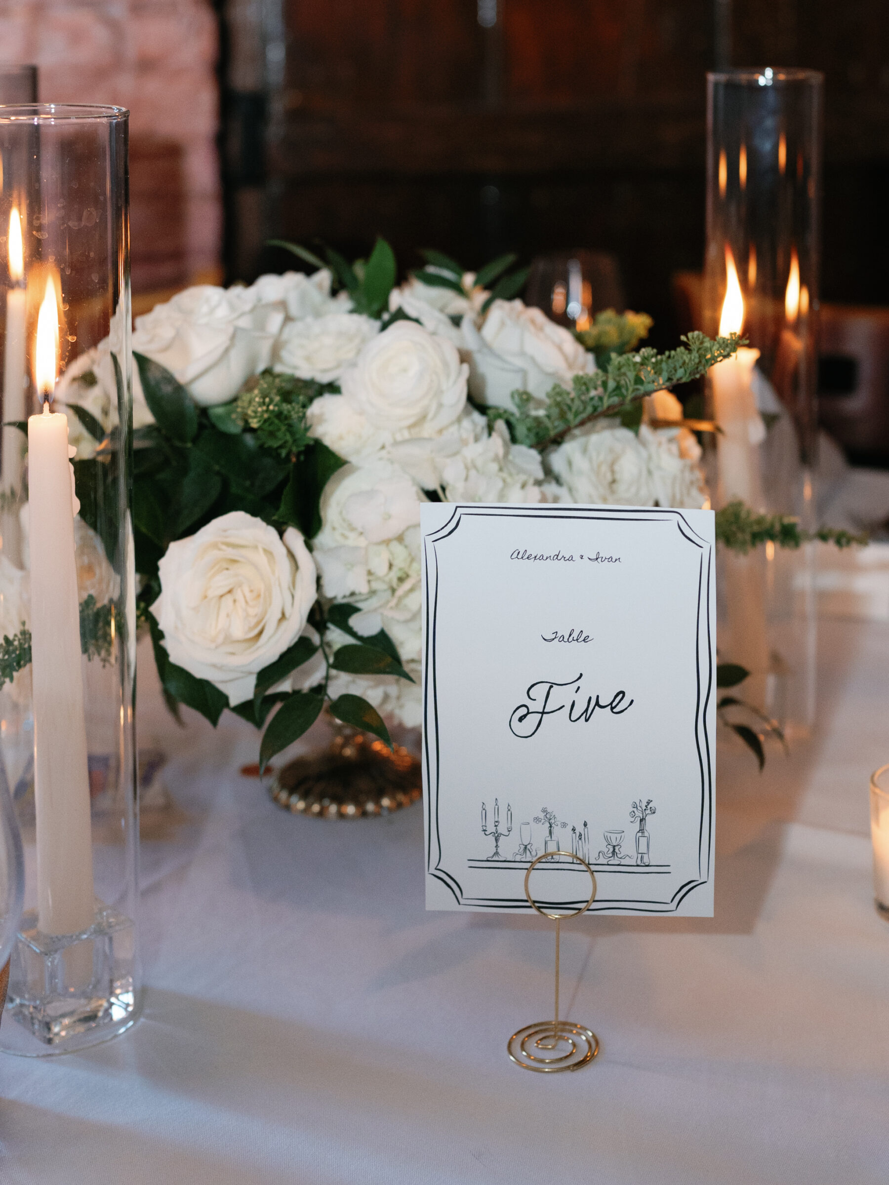 Elegant table number card reading “Five” in flowing script, set before a lush white rose centerpiece and glowing candles at an Alexandria wedding reception.