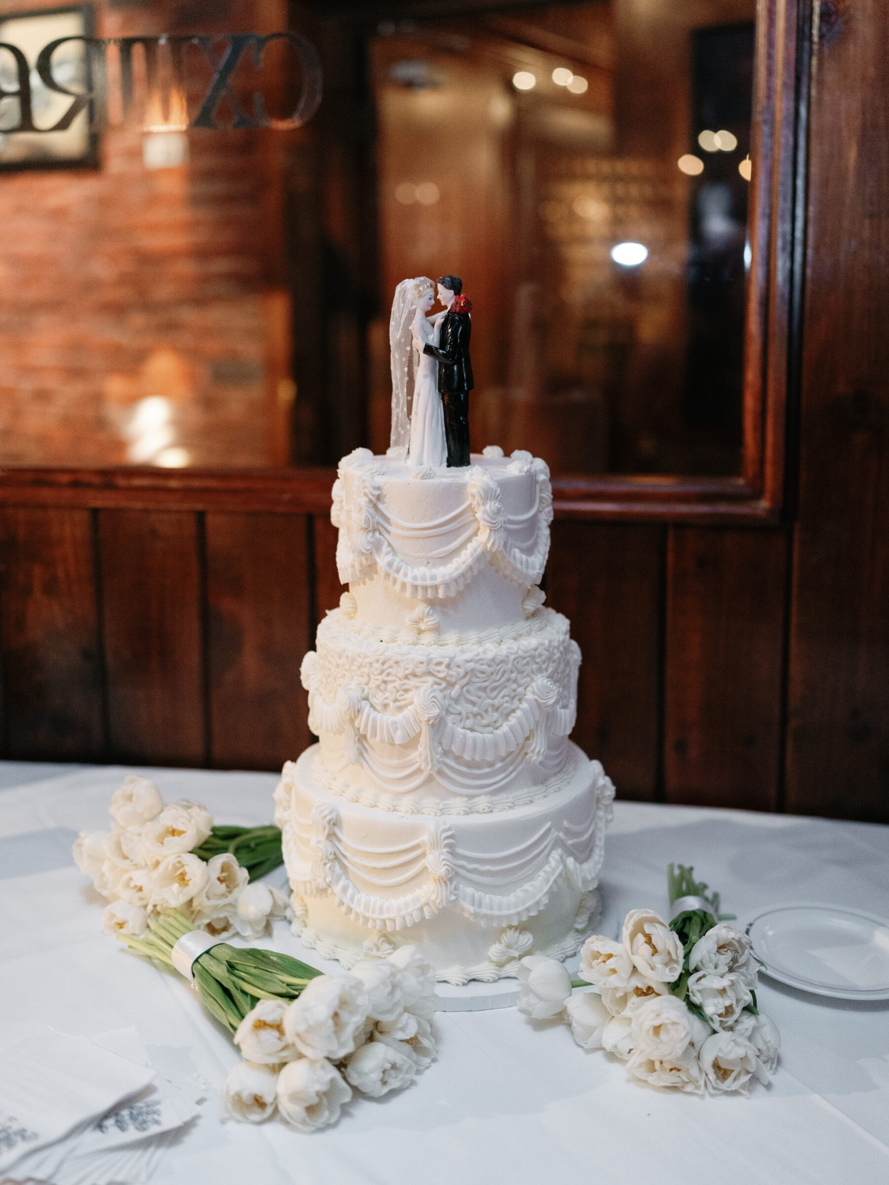 Three-tier white wedding cake with elegantly draped frosting tiers and a classic bride-and-groom topper, surrounded by loose white tulip bouquets on a linen-covered table.