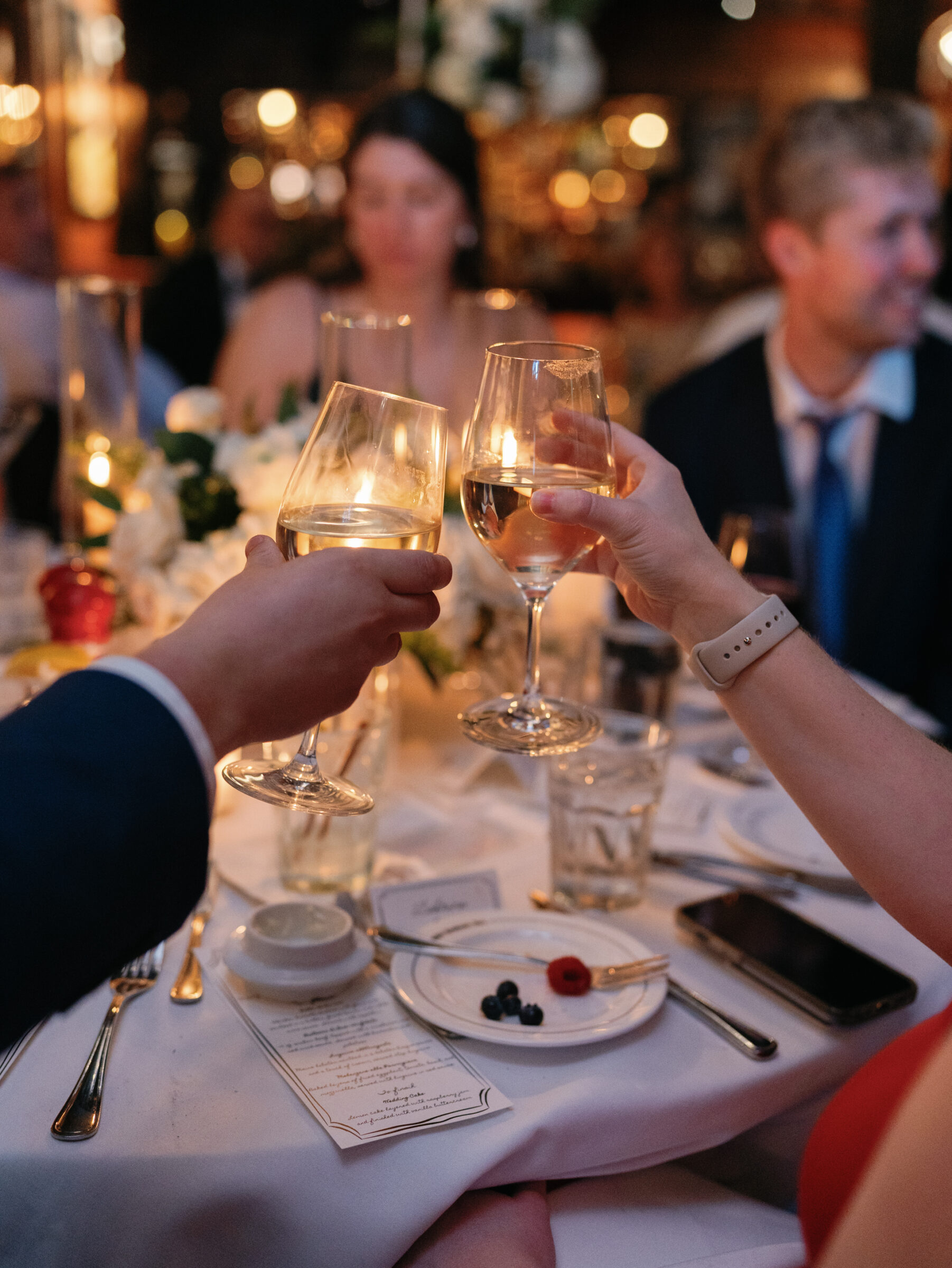 Two guests clink wine glasses over a candlelit table set with white linens, a floral centerpiece, and elegant place settings.