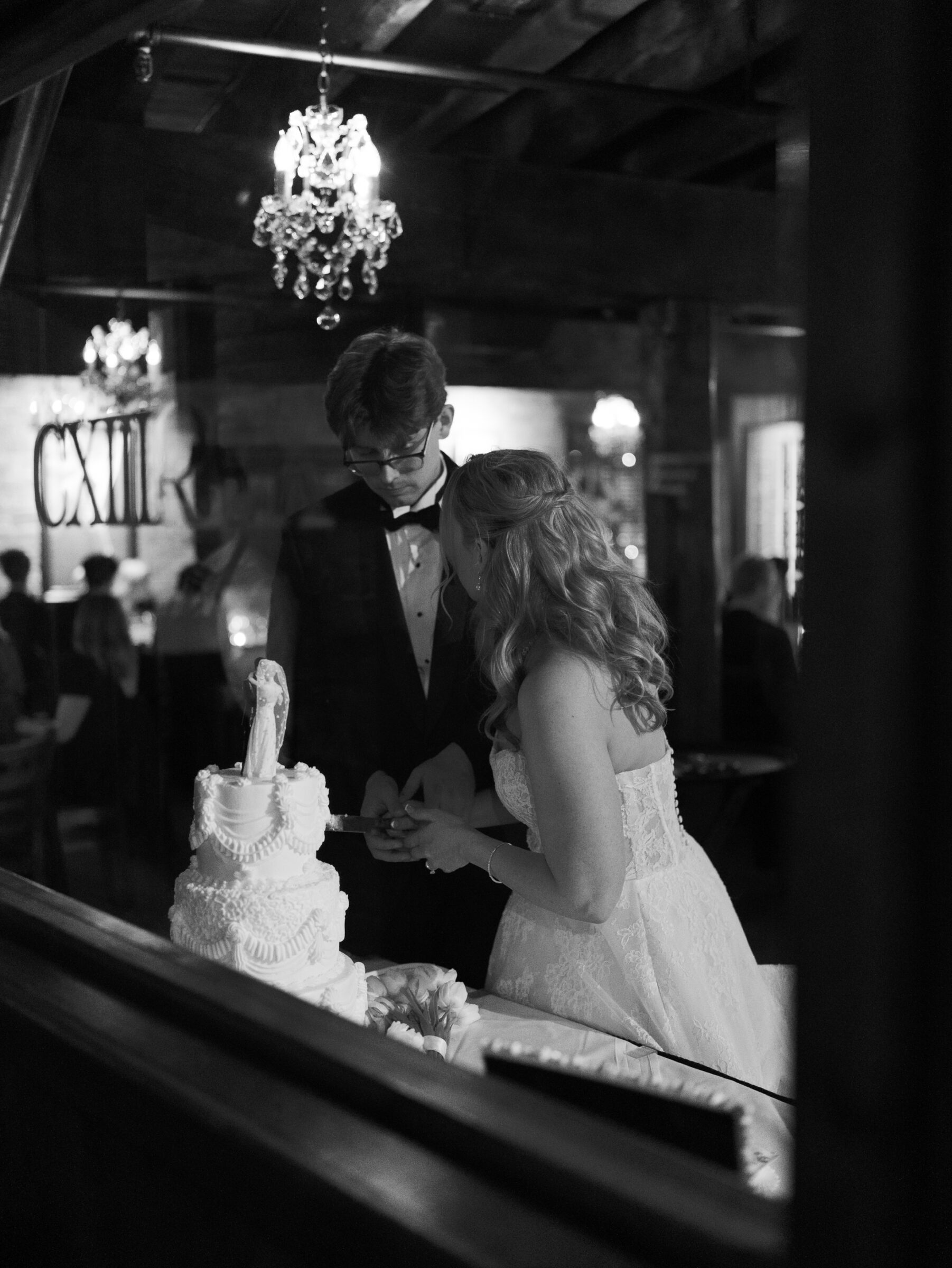 At Landini Brothers Restaurant in Old Town Alexandria, the bride and groom cut their three-tier wedding cake beneath a crystal chandelier, her lace gown and the cake’s elegant frosting framing a candlelit, intimate celebration.