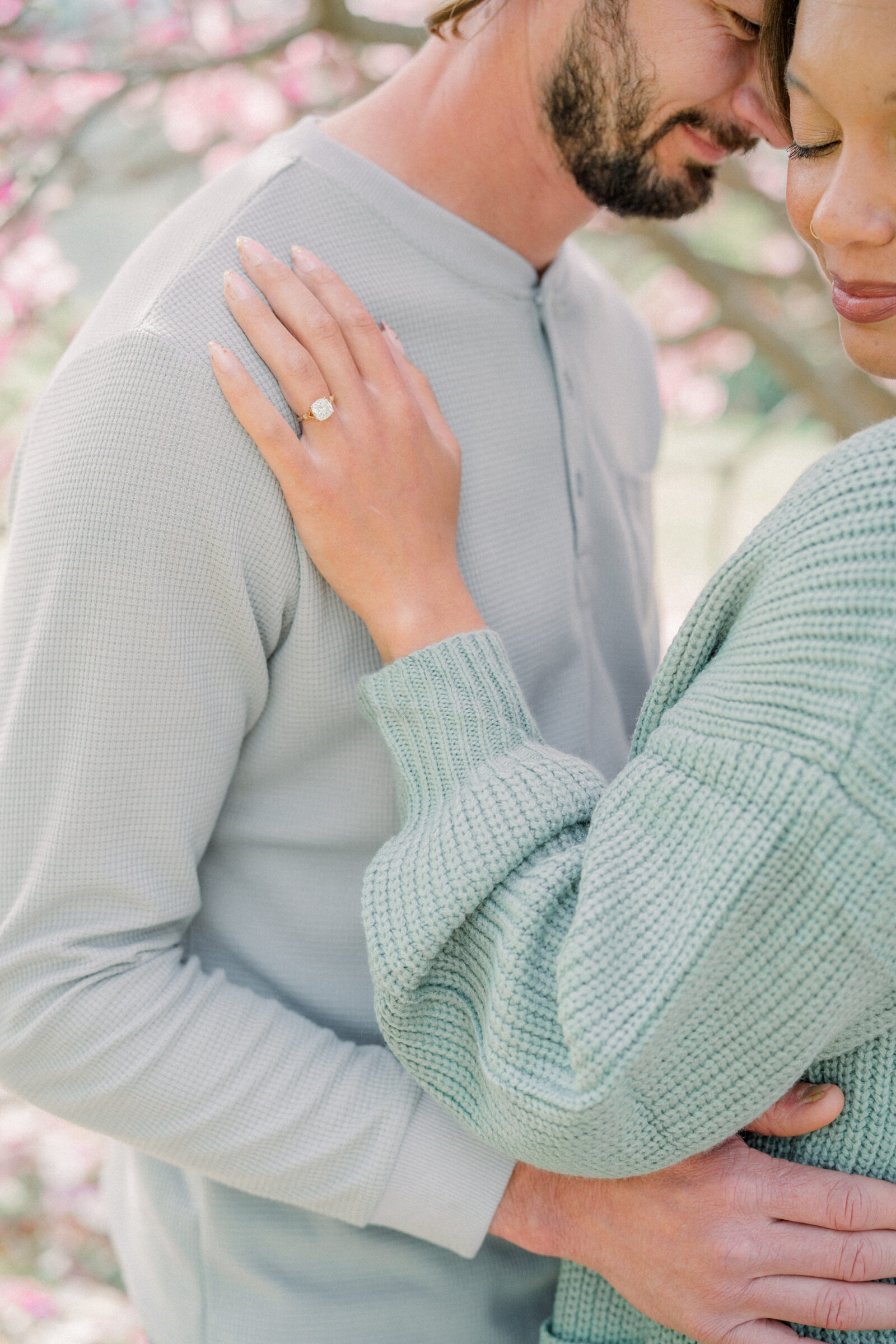 Close‑up of bride’s hand showcasing her engagement ring as she embraces her partner beneath pink magnolia blooms in a Maymont Park engagement session