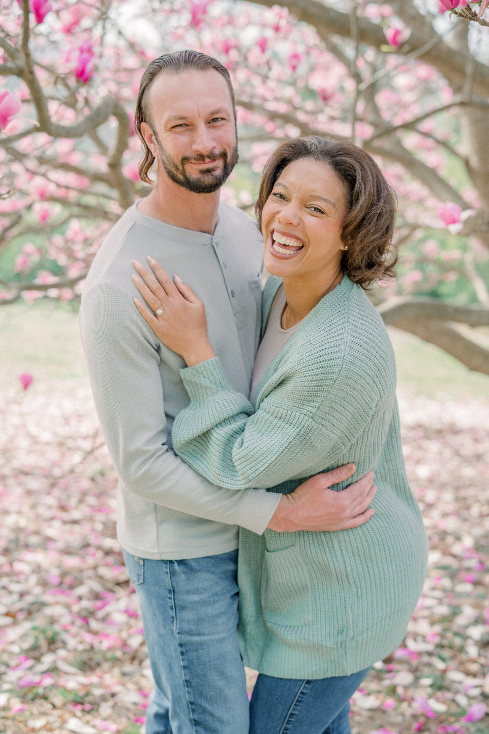 Smiling engaged couple embracing beneath a canopy of pink magnolia blossoms during their Maymont Park engagement session