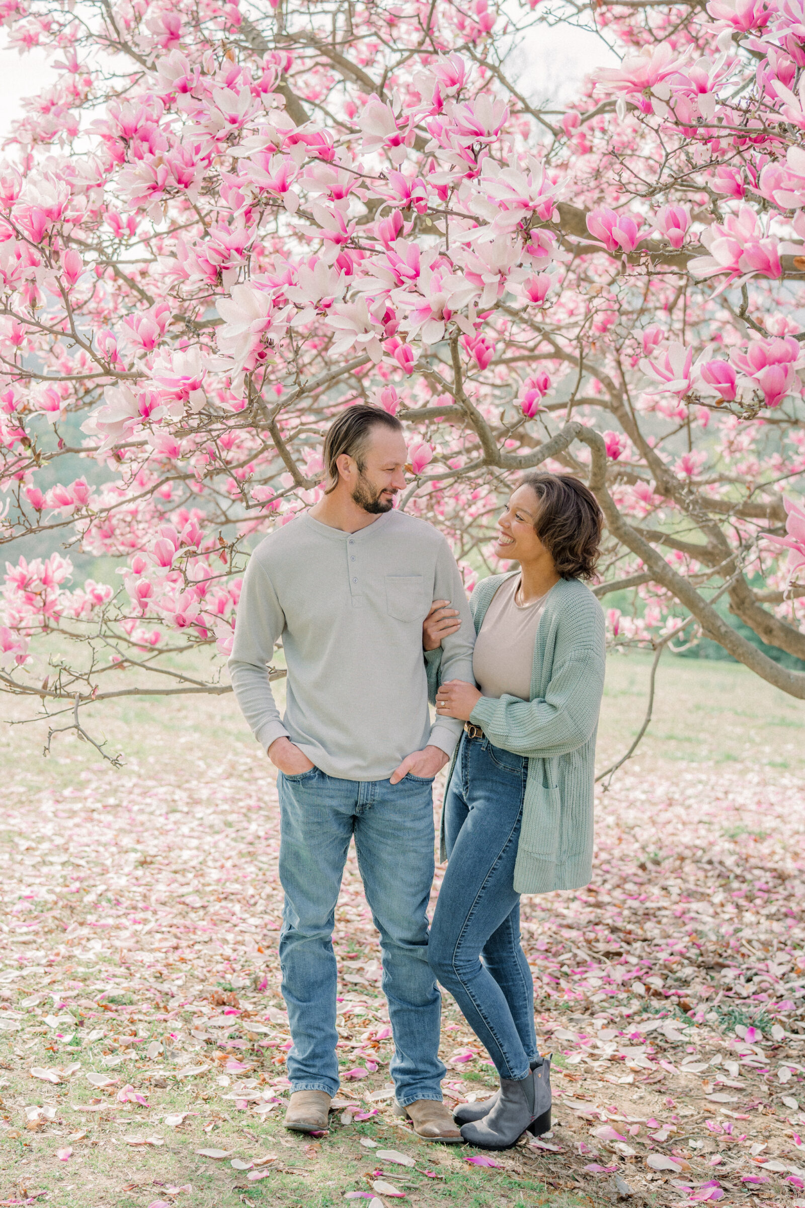Engaged couple arm‑in‑arm under a canopy of vibrant pink magnolia blossoms during their Maymont Park engagement session