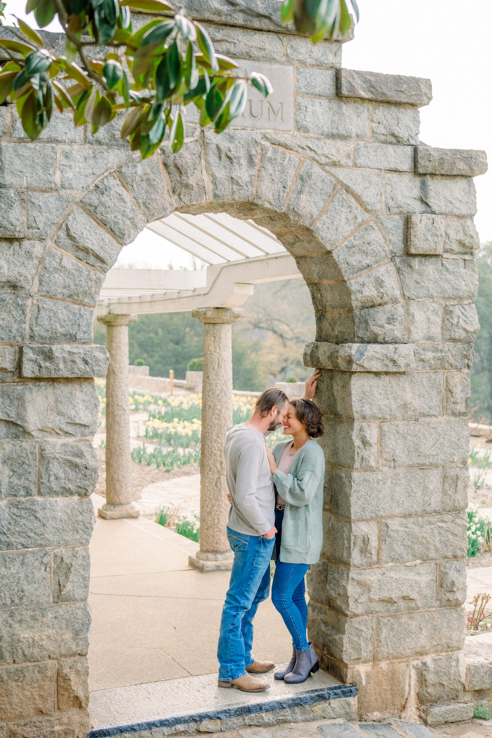 Engaged couple sharing an intimate moment beneath a rustic stone archway with blooming daffodils in the background during their Maymont Park engagement session