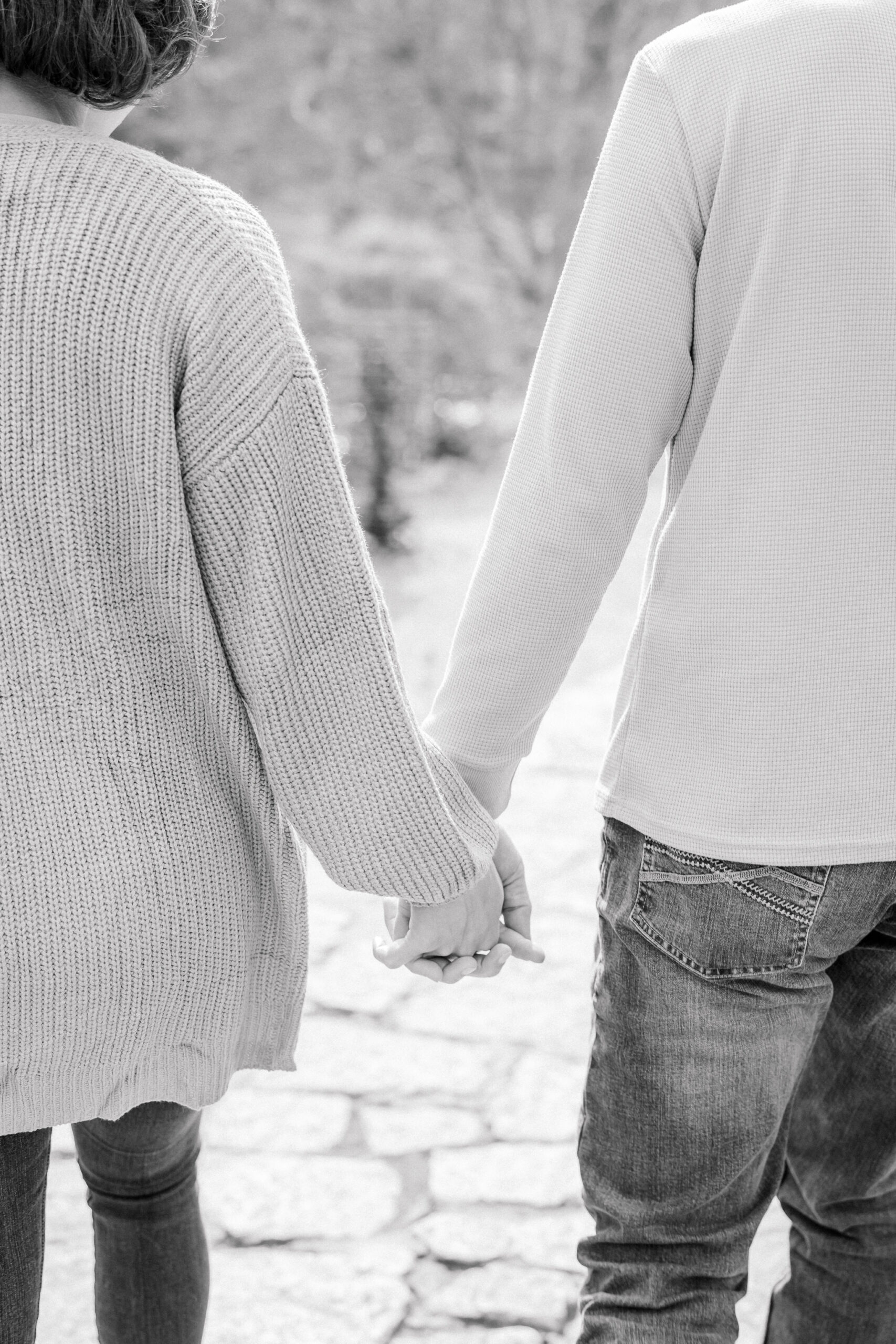 Black-and-white image showing the backs of the engaged couple as they walk away hand in hand along a stone pathway. She wears a chunky knit cardigan, he a waffle‑weave henley and jeans, their fingers intertwined in a gentle, intimate grip.