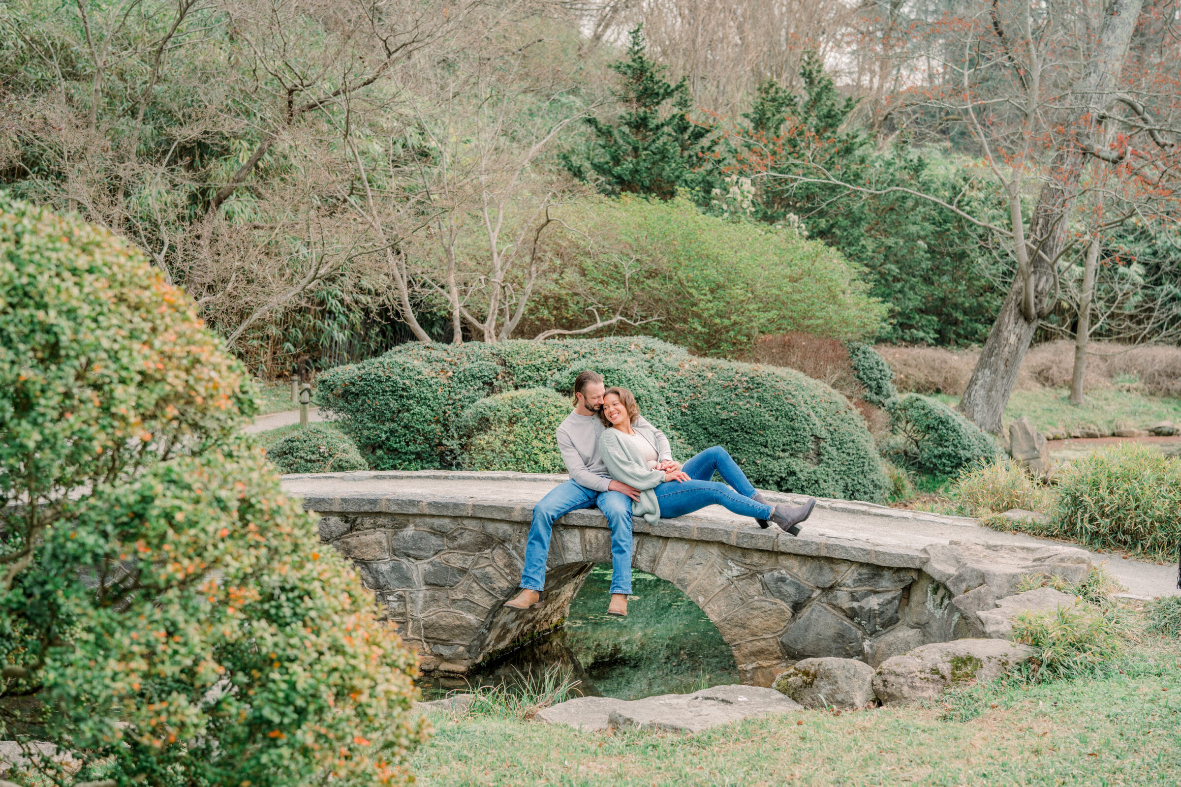 Engaged couple seated on a low stone arch bridge over a pond, she leaning back into him with a joyful smile as he wraps his arms around her, framed by landscaped shrubs and bare winter trees at Maymont Park