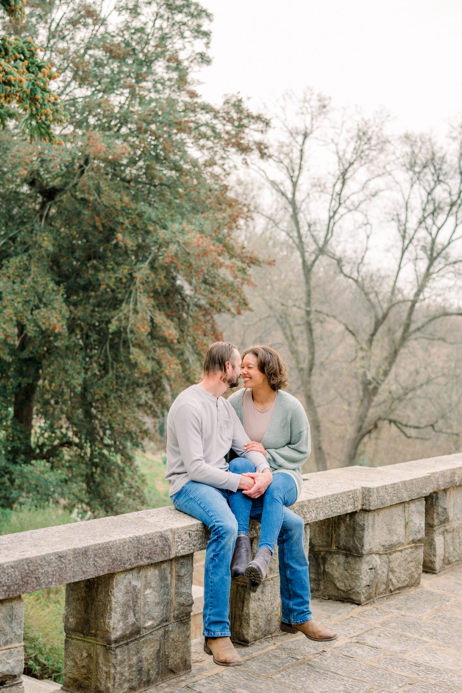 Engaged couple sitting on a wide stone ledge overlooking a garden, she perched beside him and leaning in as they touch noses, bare winter trees and evergreen shrubs framing the scene at Maymont Park.
