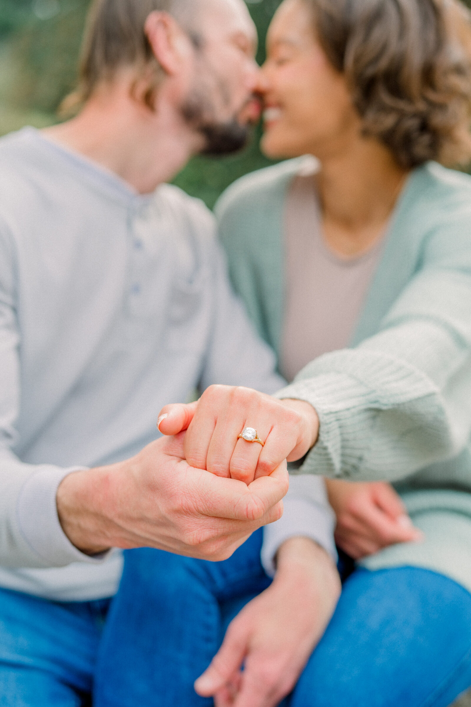 A soft-focus portrait of the engaged couple sharing a gentle nose‑touch in the garden, with her left hand—adorned with a sparkling solitaire engagement ring on a golden band—extended into the foreground and held in his hand.