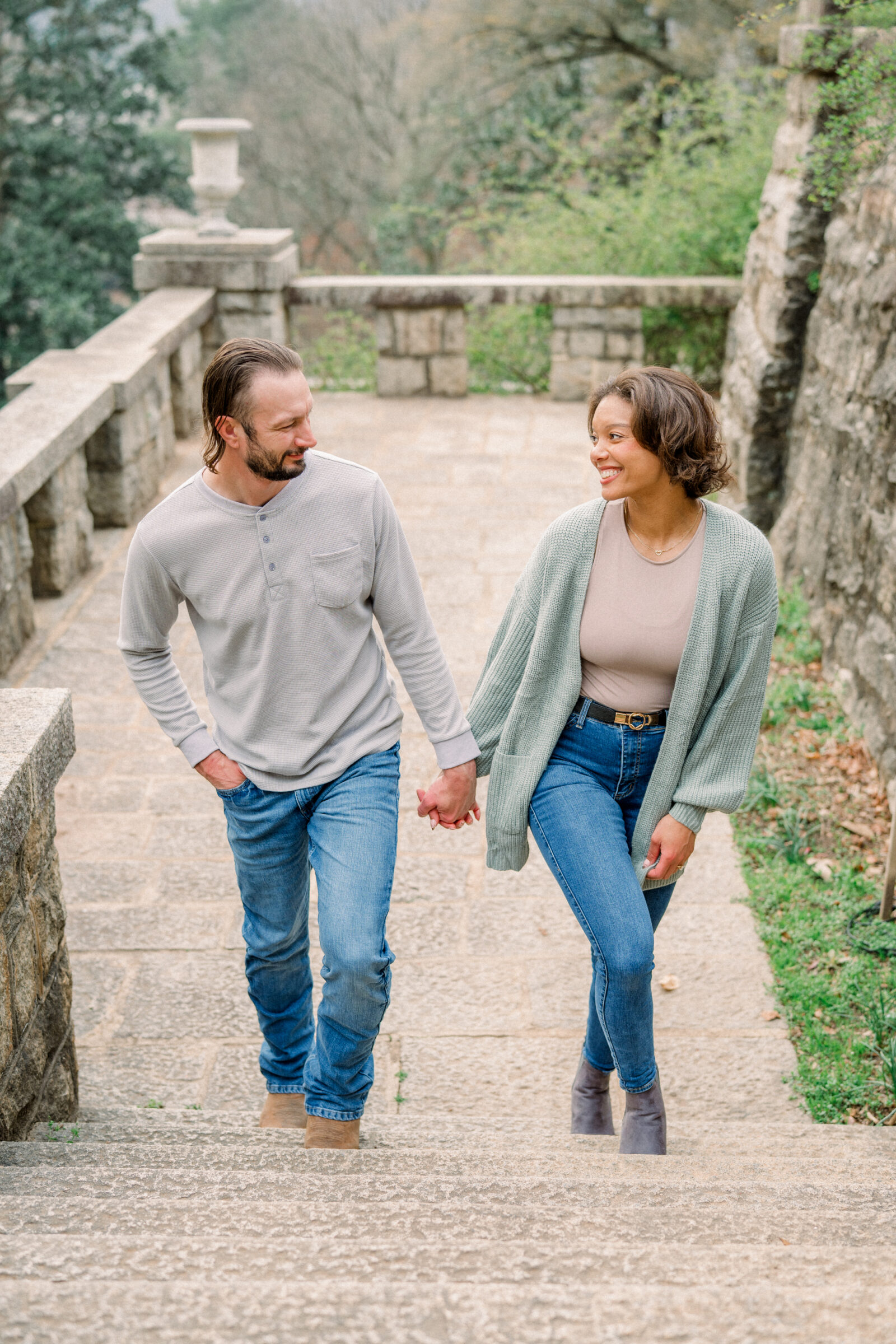A smiling couple ascends a weathered stone staircase beneath a moss‑topped balustrade. He wears a light waffle‑knit henley and jeans, she a sage green cardigan and ankle boots, their hands clasped as they look into each other’s eyes.