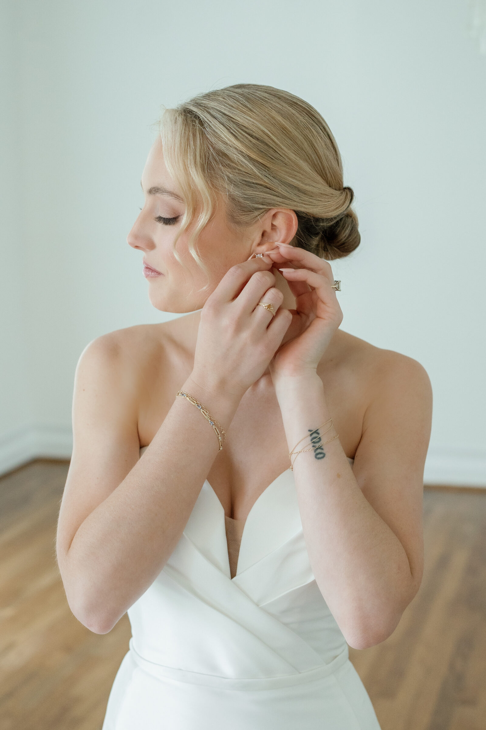 Bride getting ready inside Northfield Estate in Cumberland, VA, delicately putting on earrings while wearing a strapless satin gown—capturing an intimate and elegant bridal moment in a light-filled suite near Richmond.