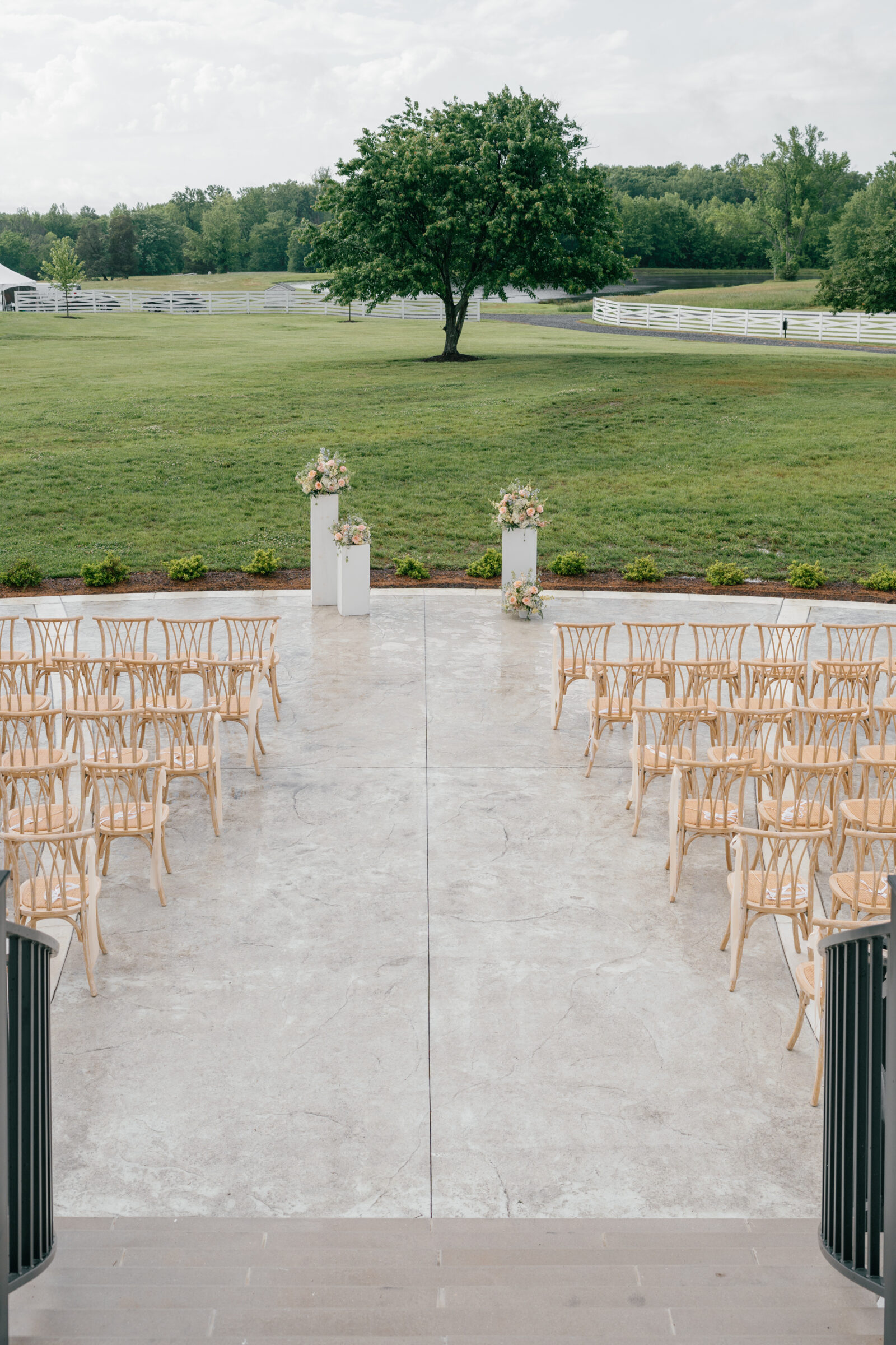 Ceremony setup on the terrace at Northfield Estate in Cumberland, VA, featuring natural wood chairs and floral pillars overlooking the estate’s rolling fields, white fencing, and signature tree—an elegant outdoor wedding ceremony space near Richmond.