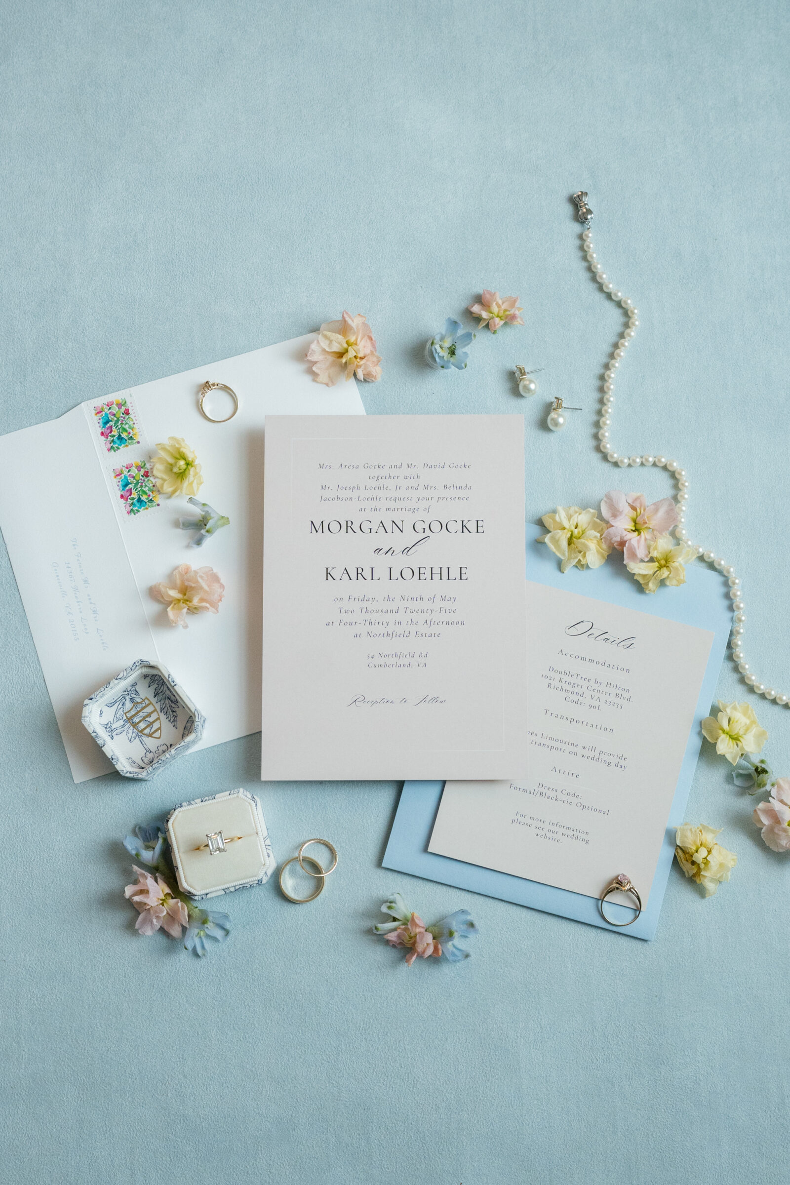 Flat lay of a wedding invitation suite for Northfield Estate wedding in Cumberland, VA, arranged on a soft blue background with cream and blue stationery, pastel spring blossoms, gold wedding bands, and a pearl necklace.