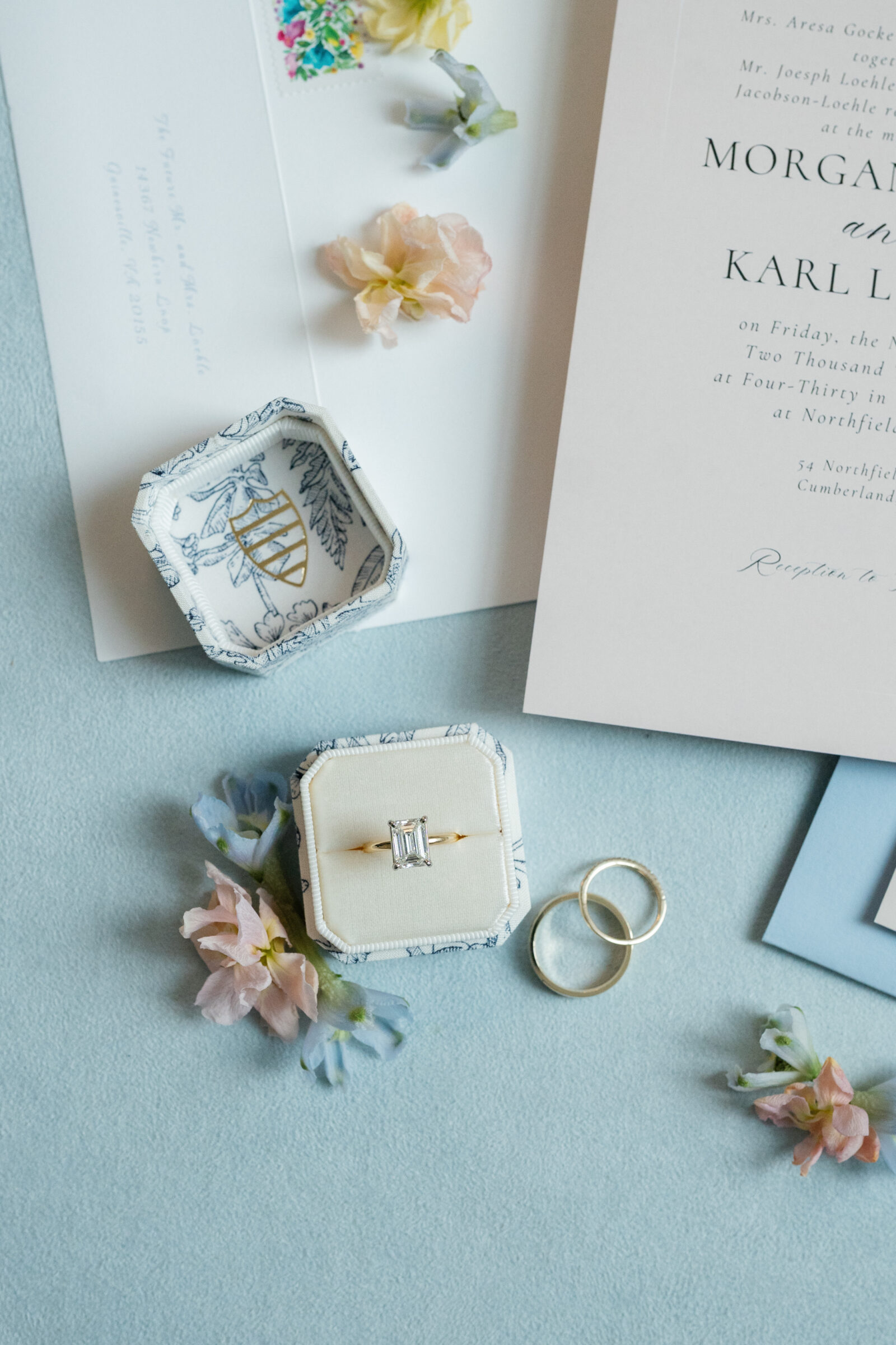 Flat lay detail of a velvet ring box holding an emerald‑cut diamond engagement ring beside two gold bands, surrounded by pastel spring blossoms and stationery on a soft blue background.