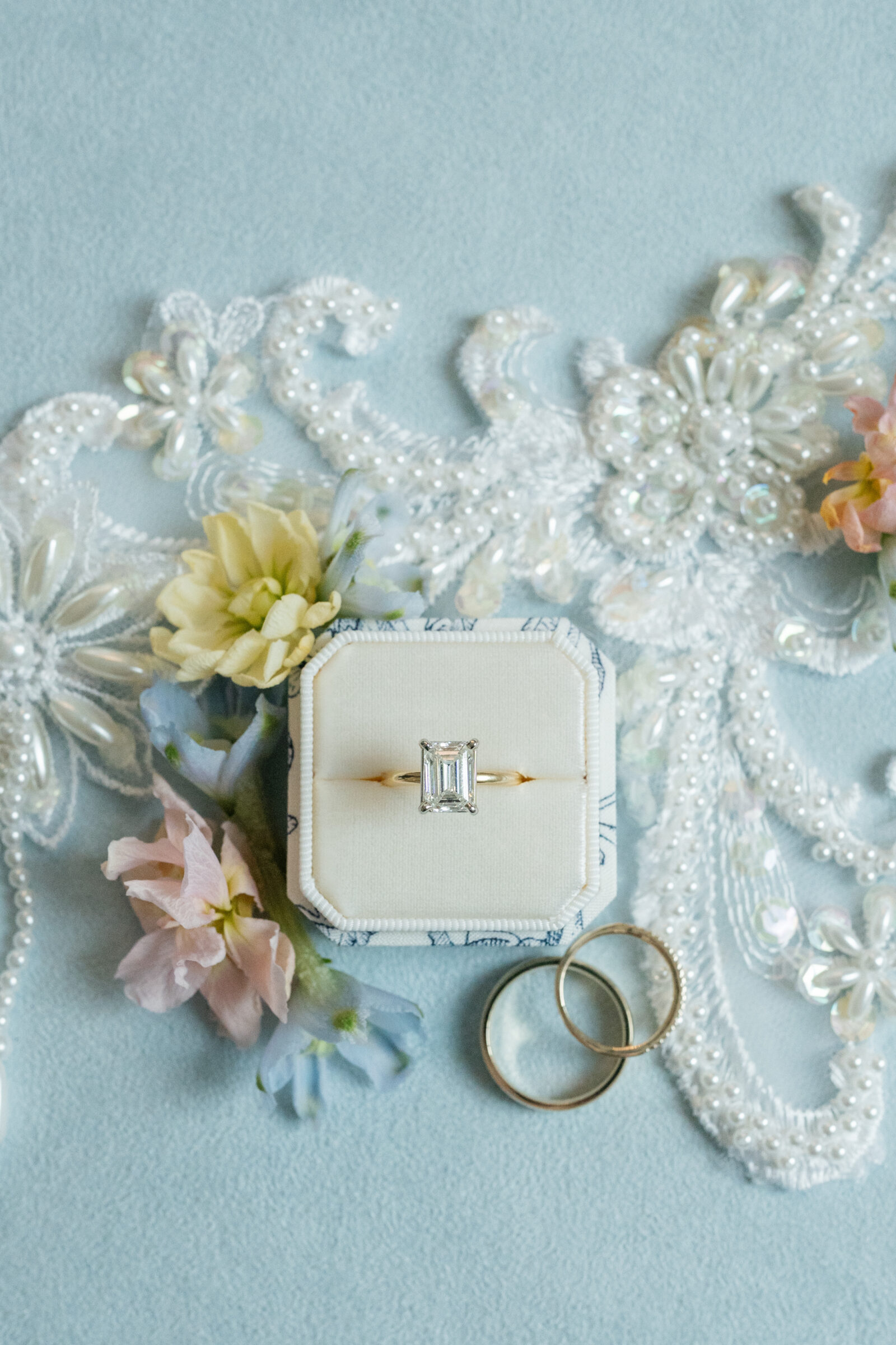 Flat lay of a velvet ring box holding an emerald‑cut diamond ring atop a beaded lace applique, flanked by two gold wedding bands and pastel spring blossoms on a soft blue background.