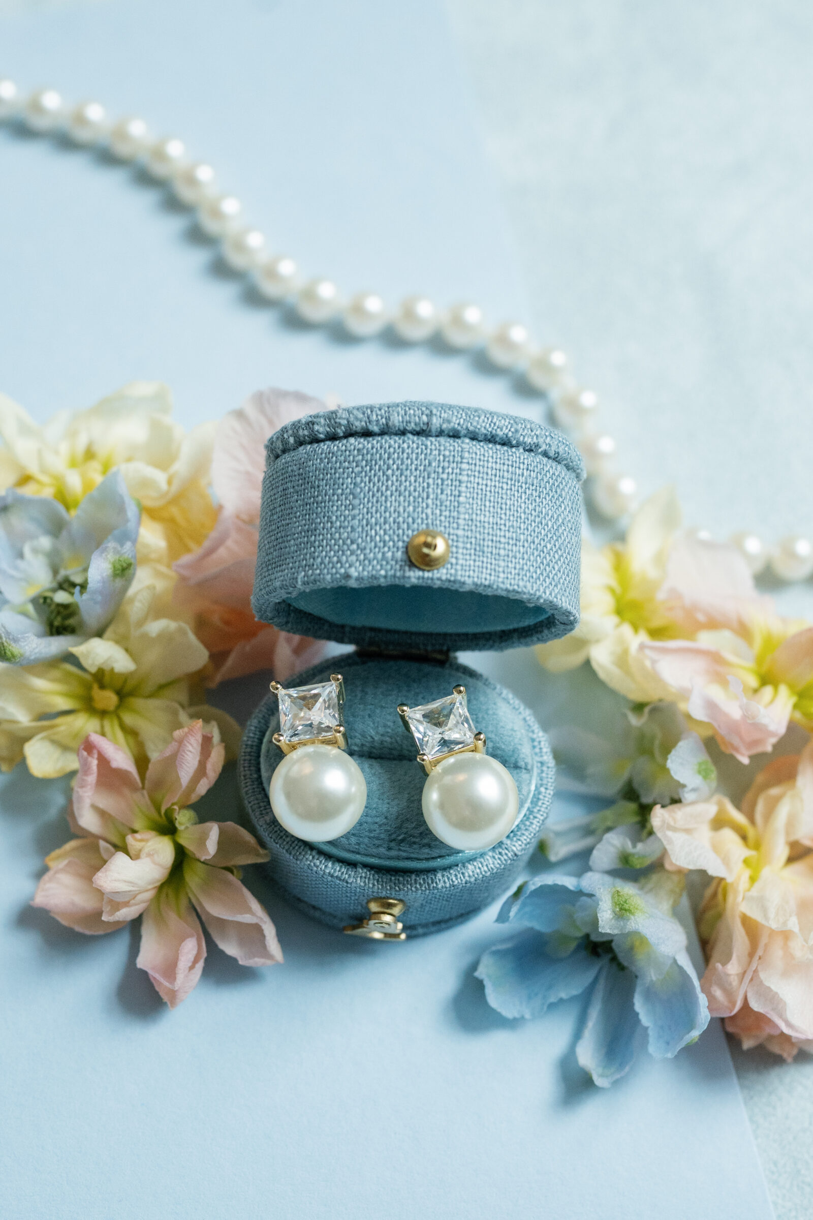 Flat lay of a light blue fabric jewelry box opened to reveal a pair of pearl drop earrings topped with square-cut crystal studs, surrounded by pastel spring blossoms and a pearl strand on a soft blue background.