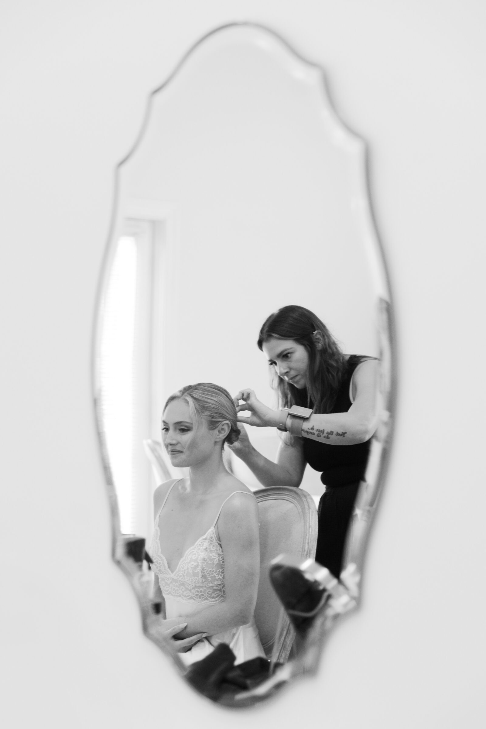 Black-and-white mirror reflection of a bride seated in a lace bodice as a stylist adjusts her updo in a softly lit room at Northfield Estate.