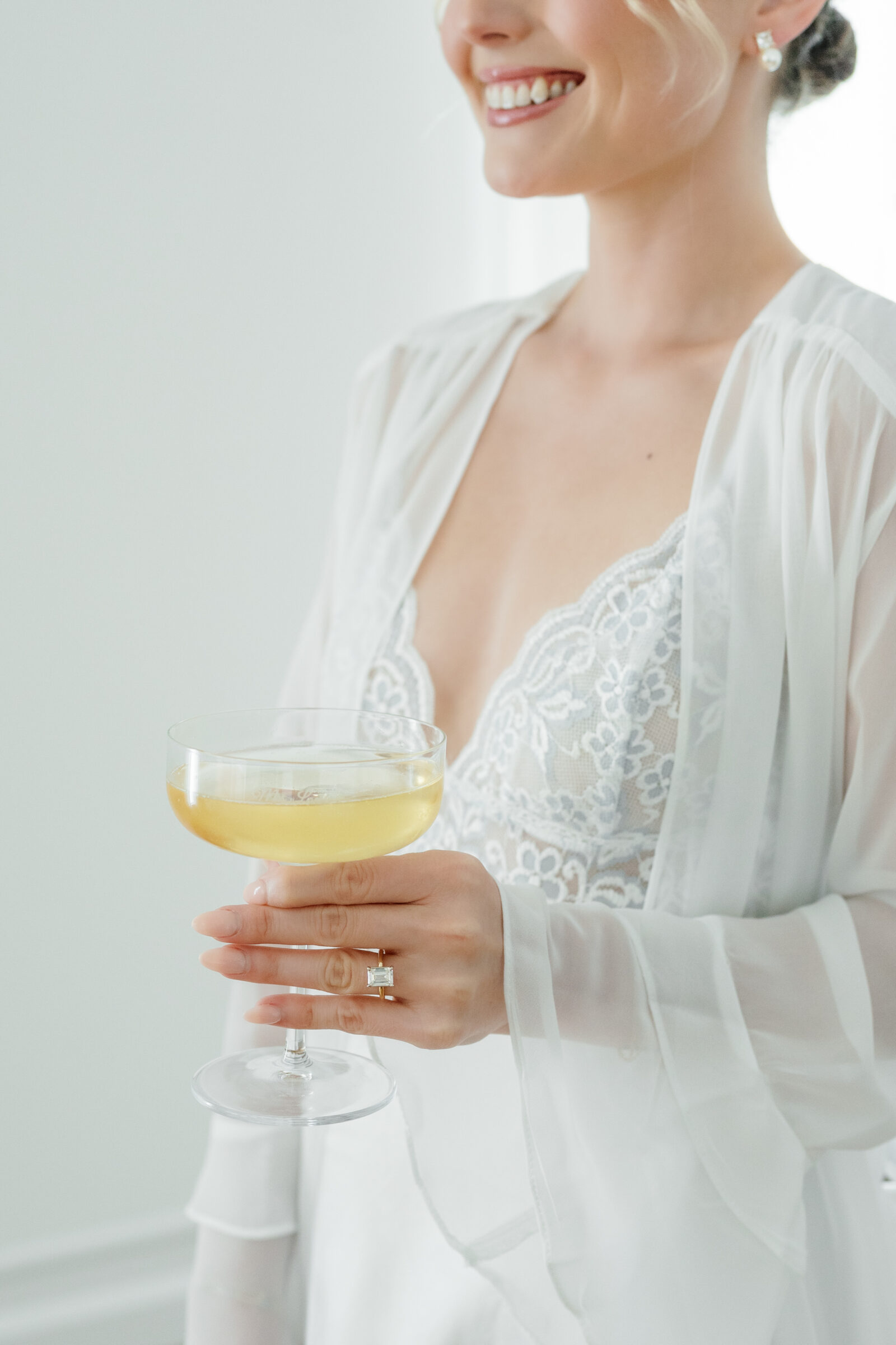 Close-up of a bride in a sheer lace robe holding a champagne coupe filled with pale yellow bubbly, with an emerald-cut engagement ring visible on her hand.