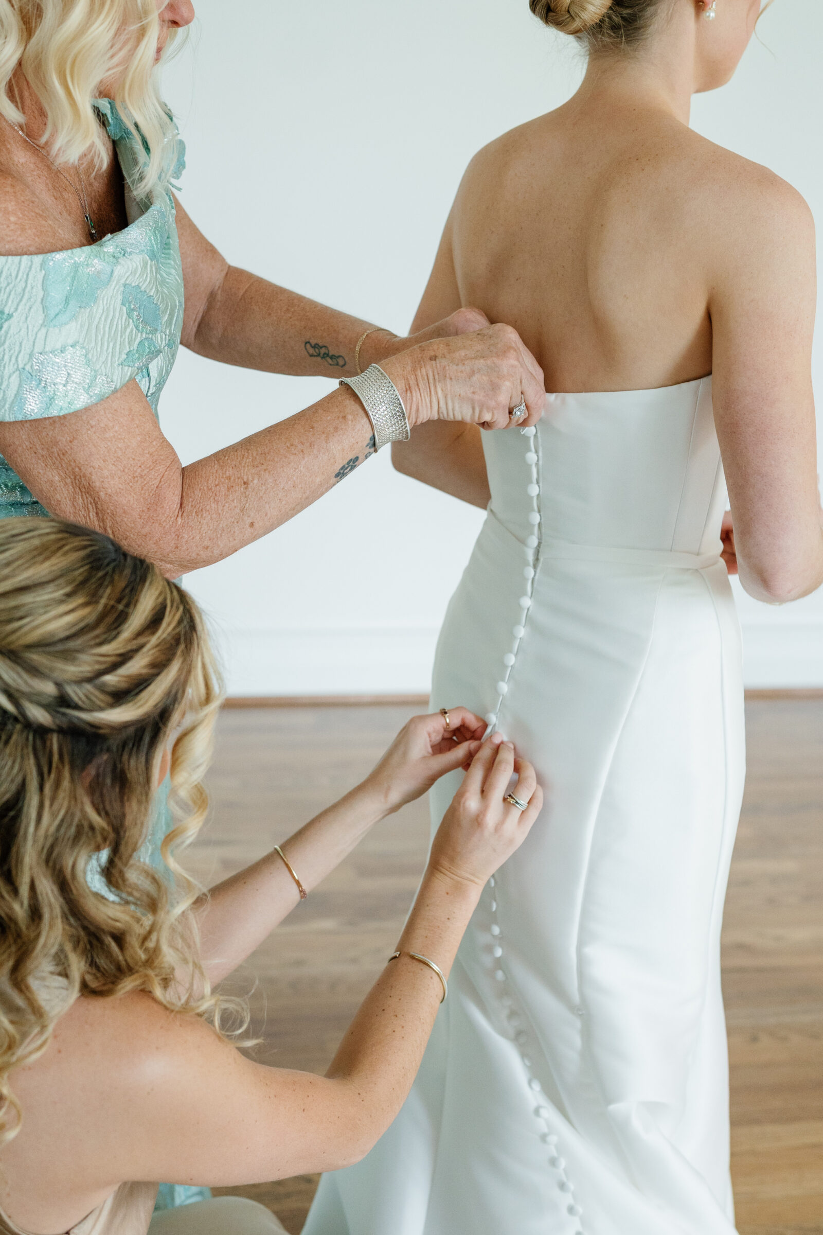 Northfield Estate Wedding - Richmond VA | A mother’s hands and a bridesmaid’s hands fastening a row of fabric-covered buttons down the back of a bride’s strapless satin gown in a bright room with hardwood floors.