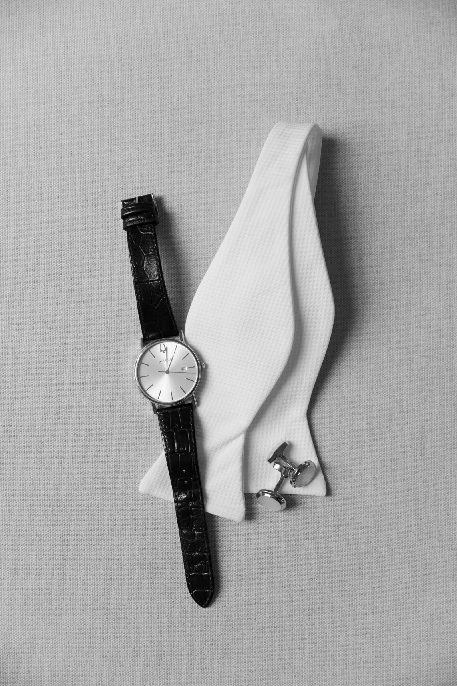 Flat lay of a groom’s accessories on a neutral fabric: a black leather‑strap wristwatch, an unknotted white textured bow tie, and a pair of silver cufflinks.