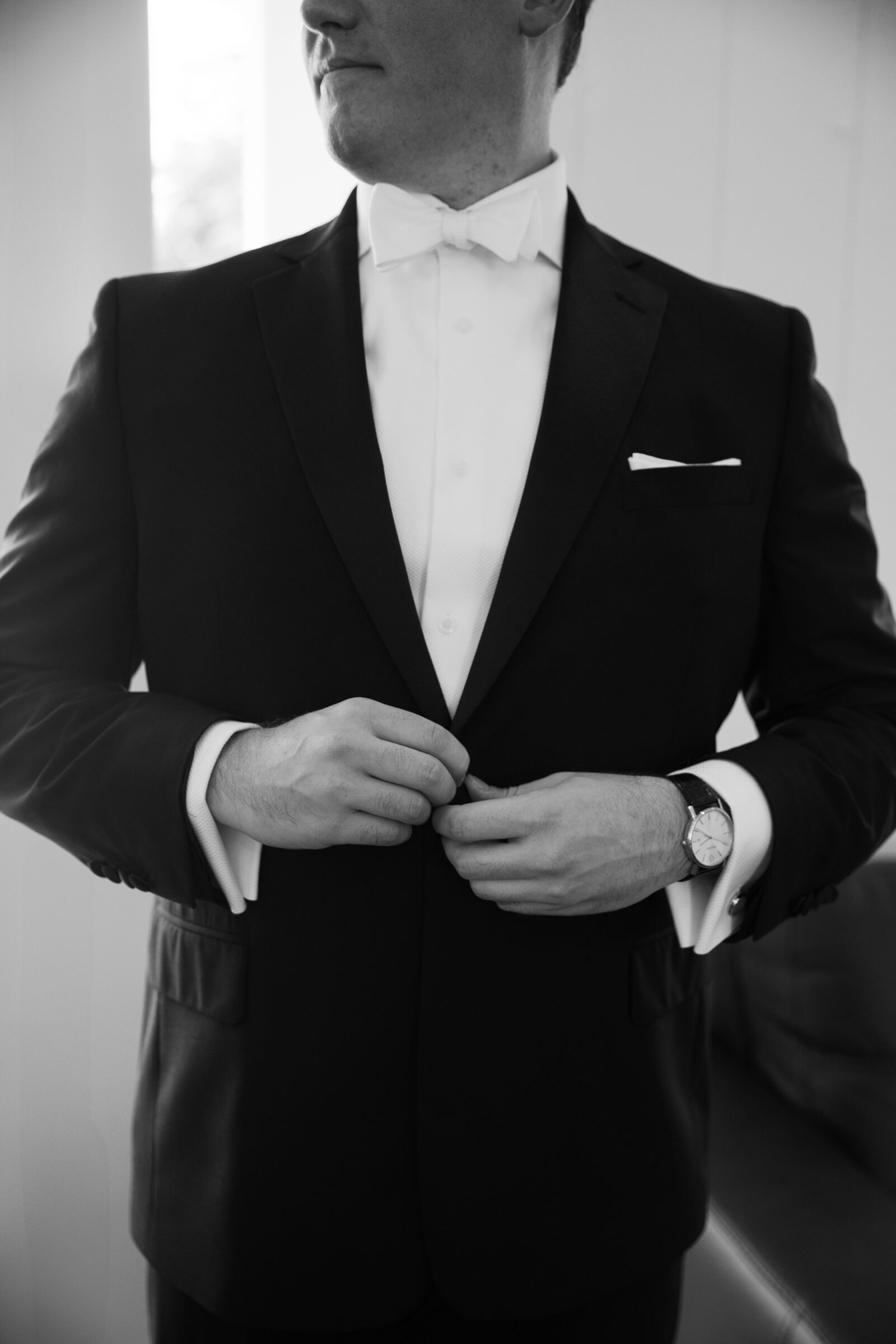 Black-and-white portrait of a groom buttoning his black tuxedo jacket over a white dress shirt with a white bow tie, his watch visible on his wrist as he prepares in a softly lit room.