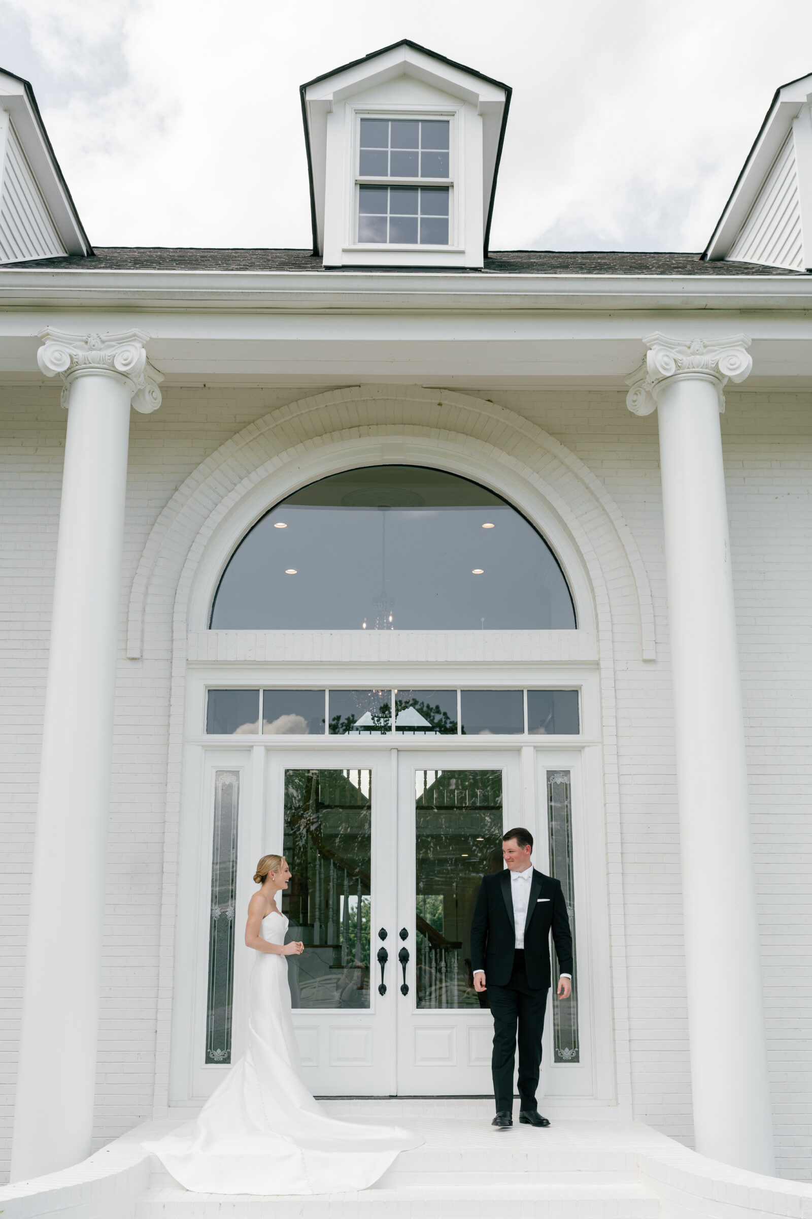 Northfield Estate Wedding - Richmond VA | Bride and groom stand on white brick steps beneath towering white columns in front of an arched glass entryway; she wears a strapless satin gown with a long train and he wears a classic black tuxedo as they face each other.