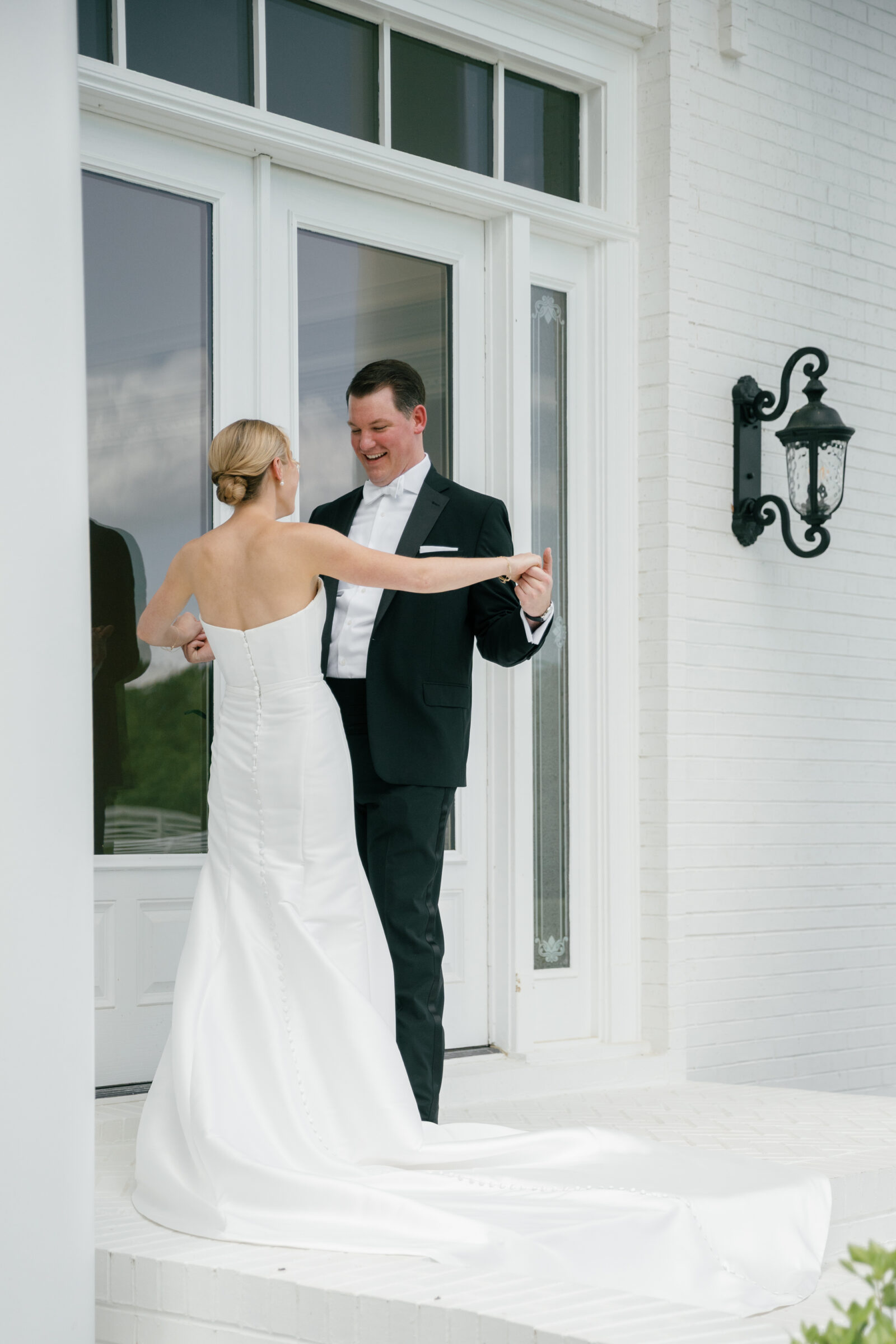 Northfield Estate Wedding - Richmond VA | Bride and groom holding hands and sharing a joyful moment on white brick steps in front of tall glass doors framed by white columns; she wears a strapless satin gown with a long buttoned train, and he wears a classic black tuxedo with a white bow tie.