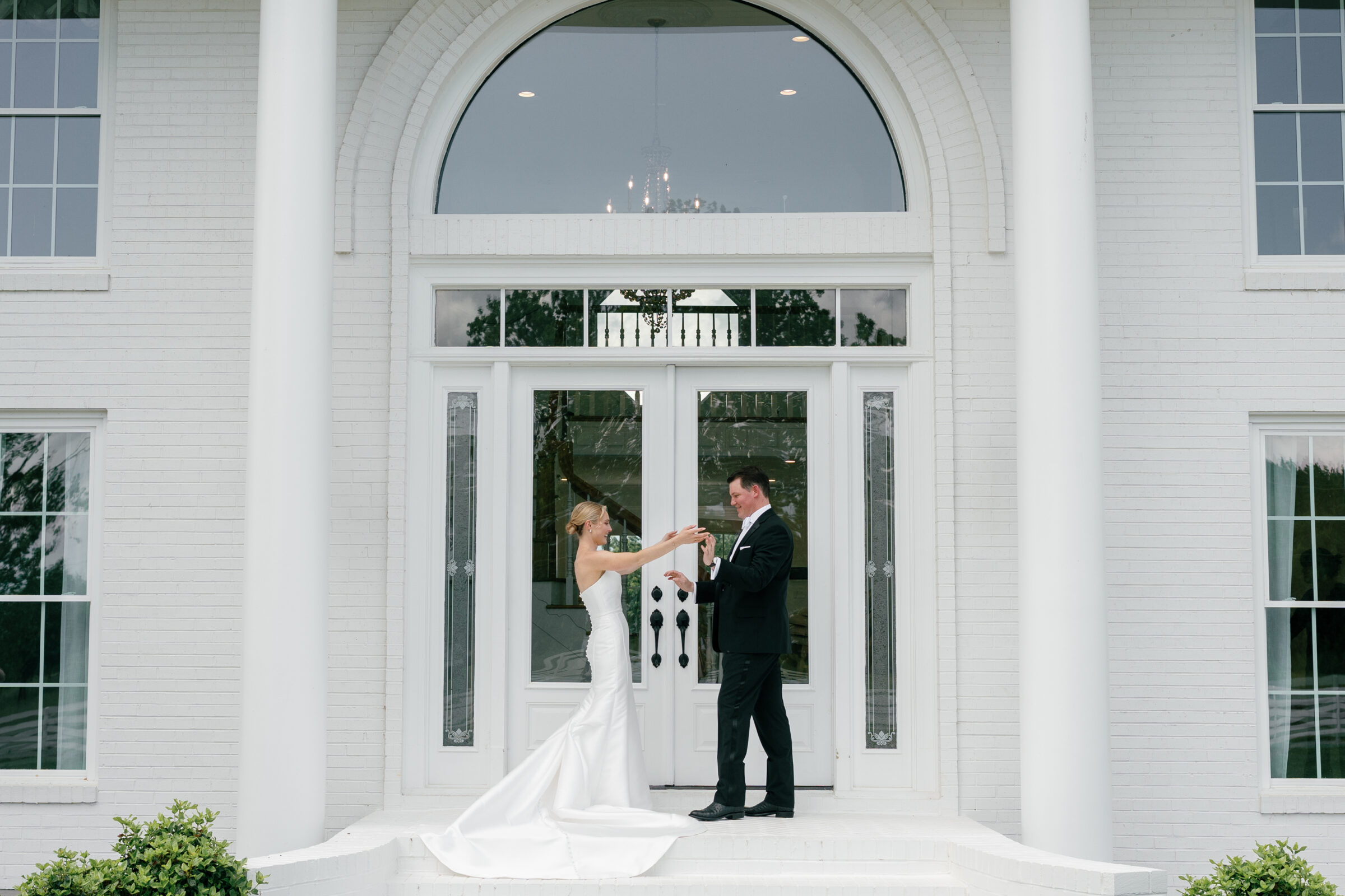 Northfield Estate Wedding - Richmond VA | Bride and groom holding hands on white brick steps in front of tall glass doors framed by white columns and an arched transom window, she in a strapless satin gown with a long buttoned train and he in a black tuxedo with a white bow tie.