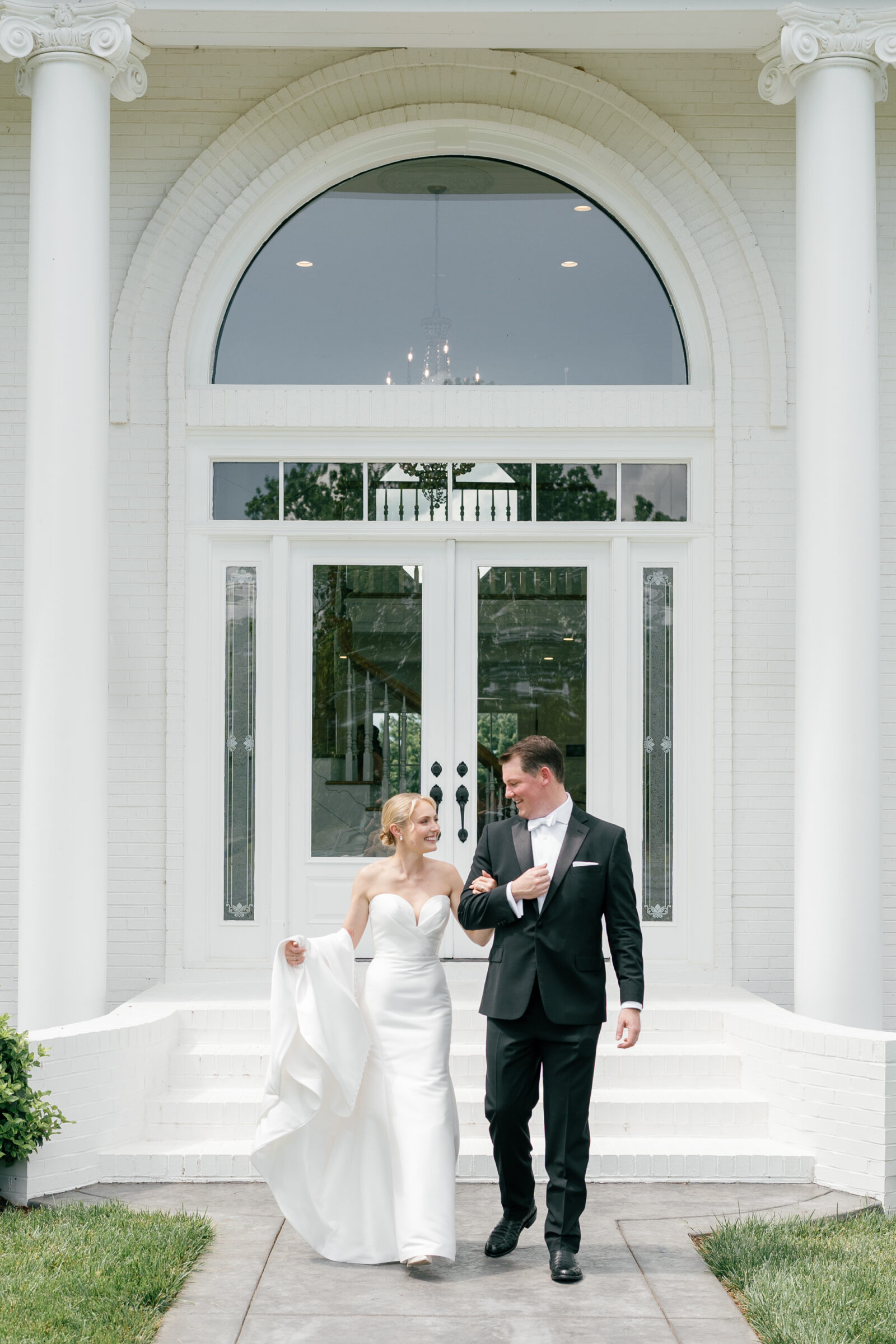 Northfield Estate Wedding - Richmond VA | Bride and groom walk arm‑in‑arm down white brick steps of a columned estate entrance, she holding her strapless satin train and smiling up at him as they step onto a stone pathway.