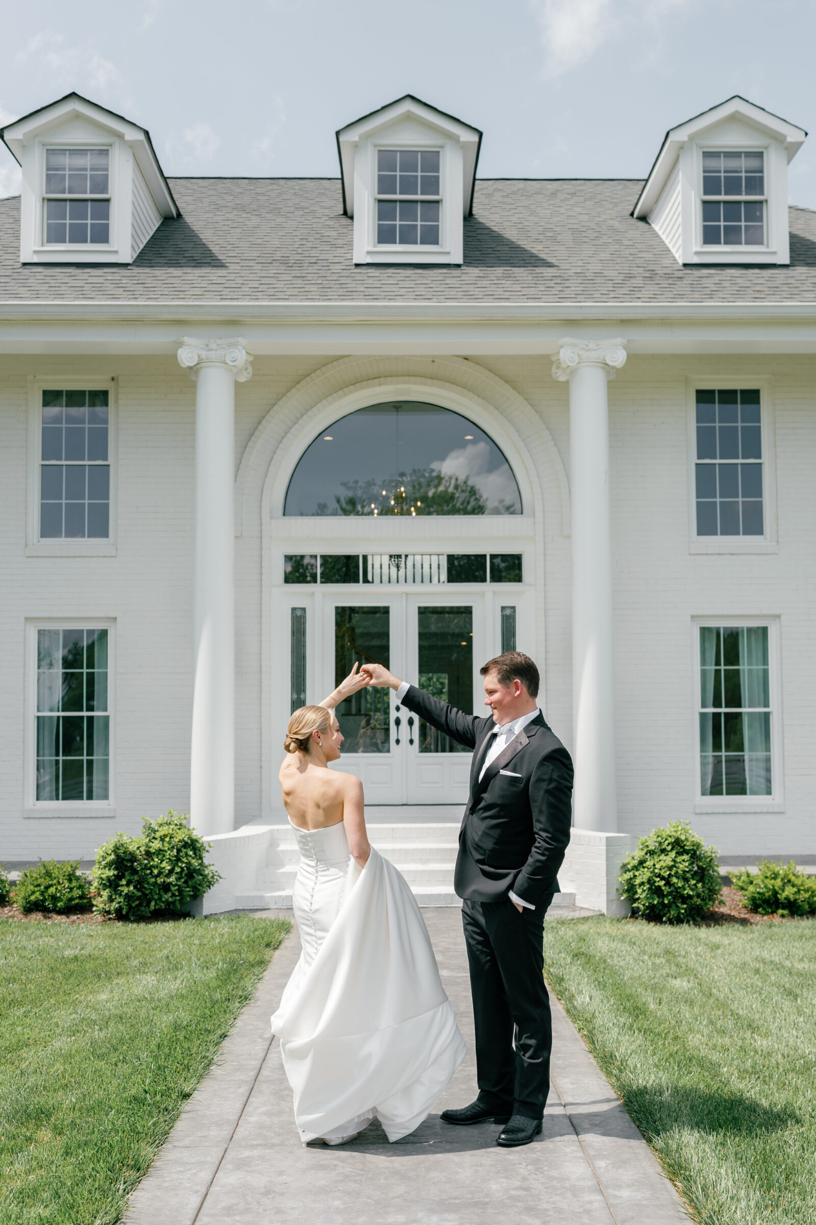 Northfield Estate Wedding - Richmond VA | Bride and groom holding hands and sharing a joyful twirl on white brick steps beneath towering white columns and an arched transom window, she in a strapless satin gown lifting her long train and he in a classic black tuxedo.