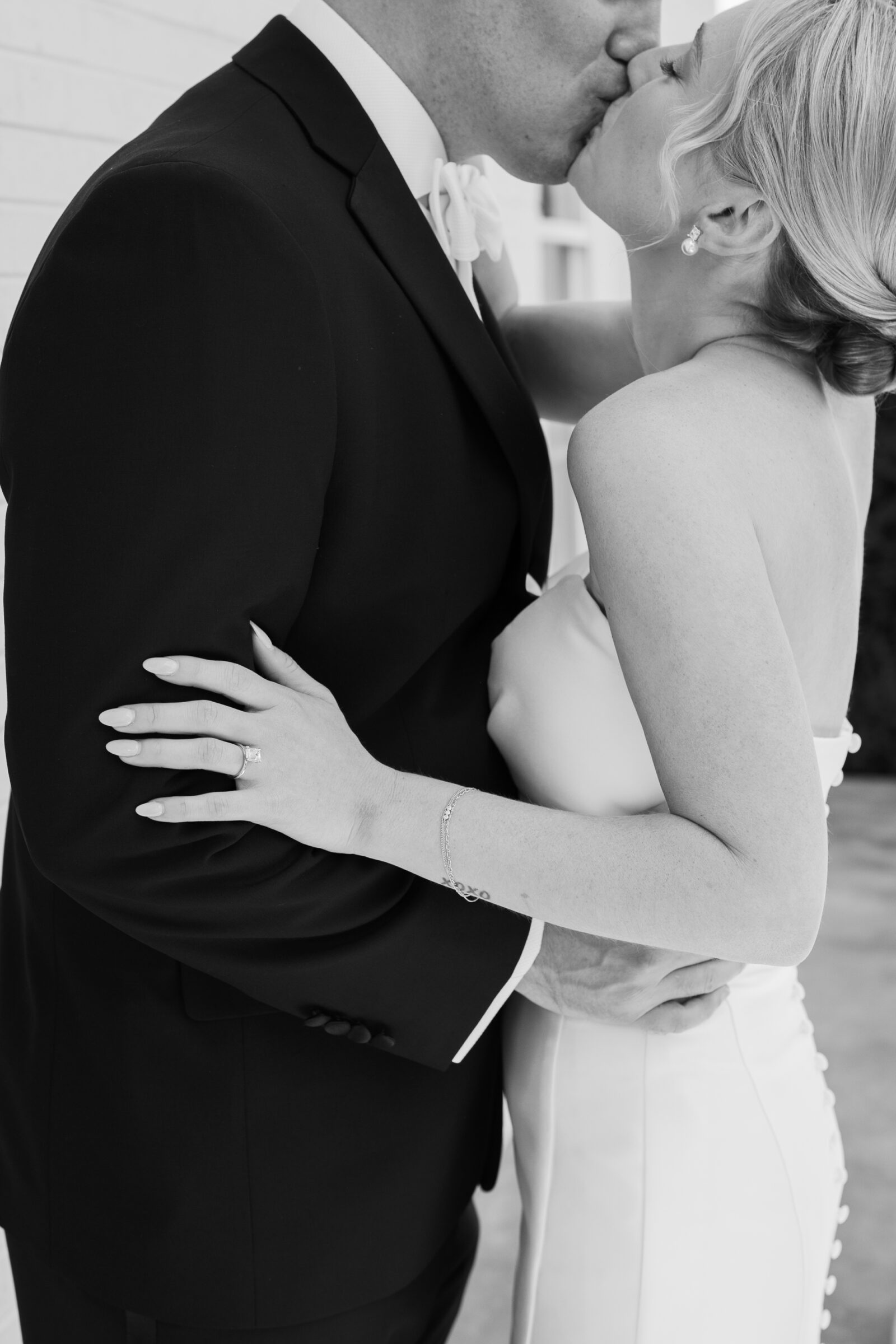 Northfield Estate Wedding - Richmond VA | Groom in a black tuxedo gently kissing the bride’s forehead as she smiles with her eyes closed, standing between a white brick wall and a stately white column, her arm wrapped around his waist.