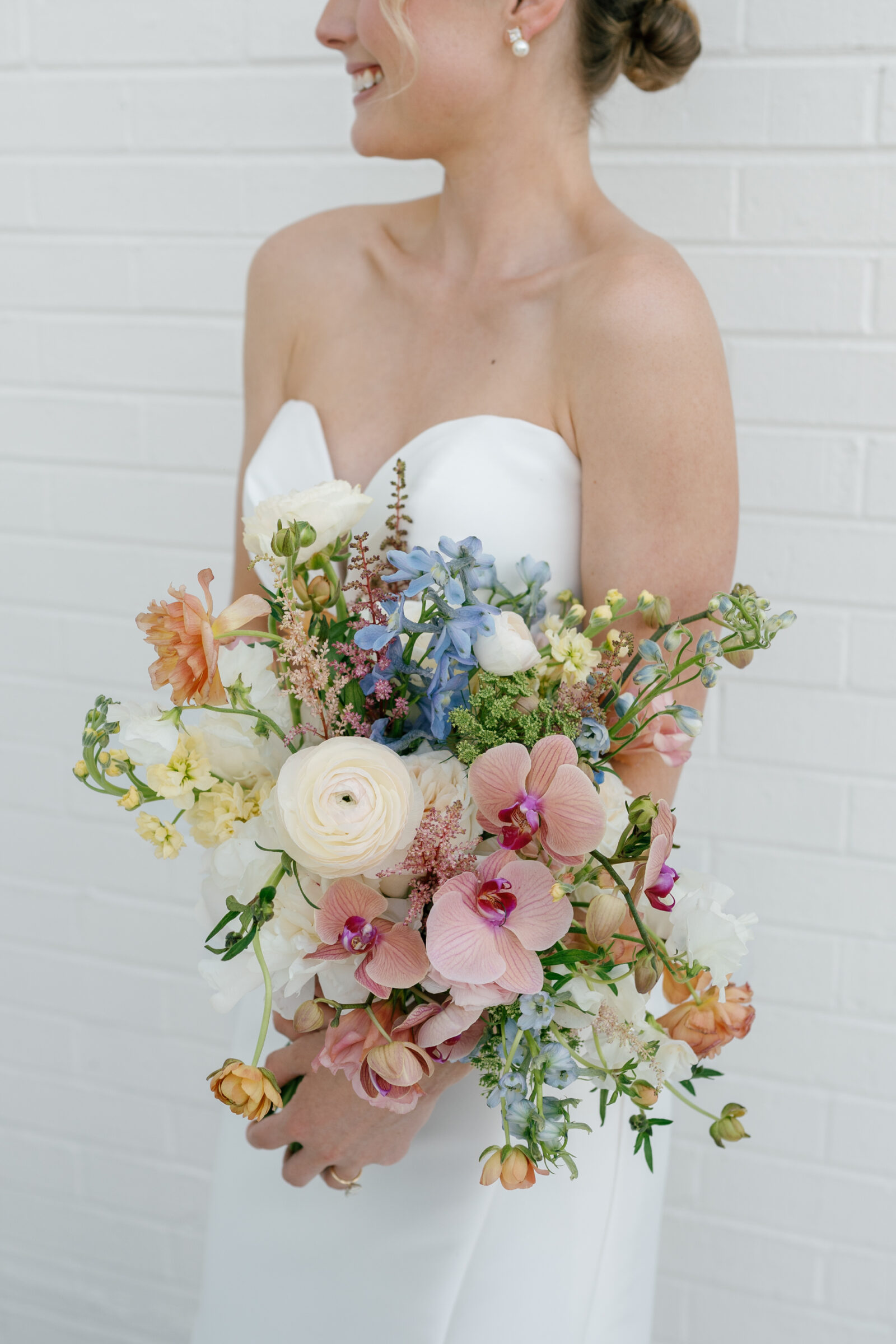 Northfield Estate Wedding - Richmond VA | Close-up of the bride’s hands clasping a loose, garden-style bouquet with pink orchids, creamy ranunculus, pale blue delphinium, peach ranunculus, and trailing greenery, set against her strapless satin gown and a white brick backdrop.